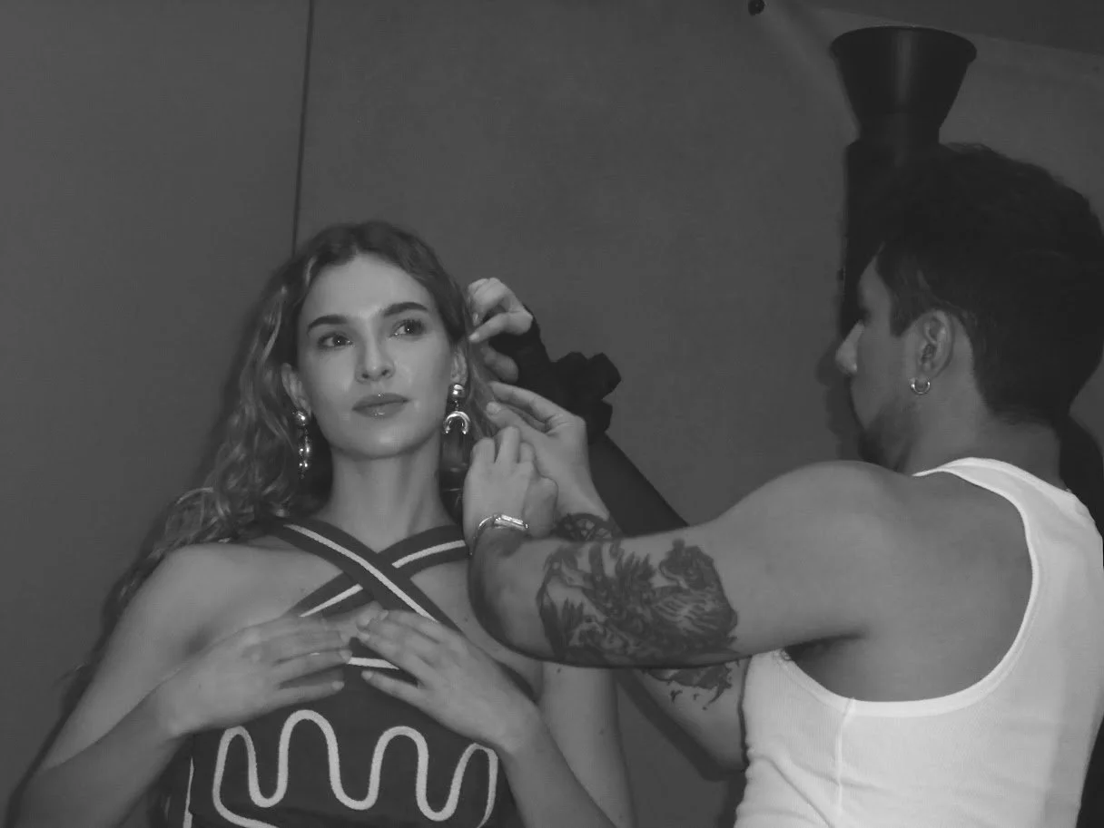 A woman with wavy hair and earrings is getting her makeup done by a makeup artist with Tattoos on his arm. The woman is wearing a top with a graphic design.