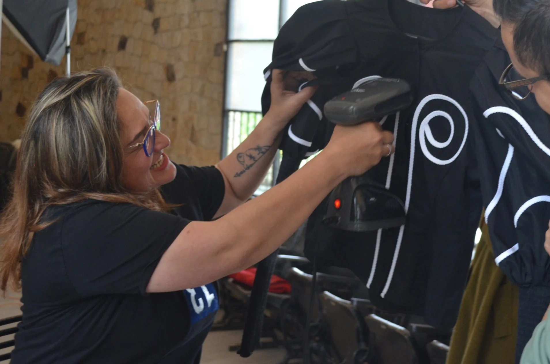 A woman with glasses and long hair helping a person in a black jacket with white trim to put on a helmet in a room with brick walls and large windows.