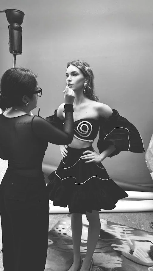 A fashion model getting her makeup or hair done by a stylist during a photoshoot in a studio, with a plain background and studio lighting.