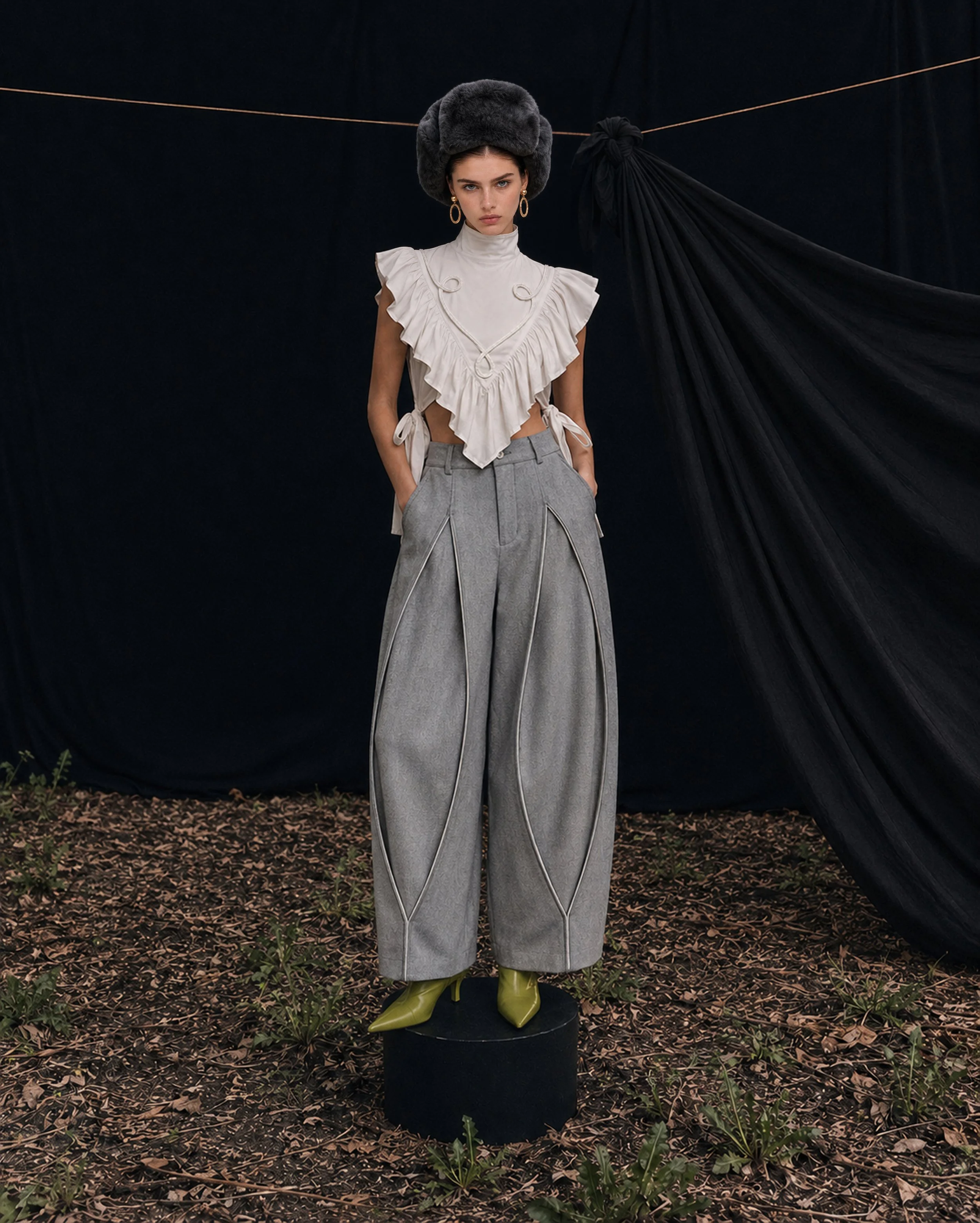 A fashion mannequin dressed in a vintage style. The mannequin wears a white ruffled top with a high neckline, tied at the sides, gray high-waisted wide-leg pants with black piping, green pointed high heels, and a large gray furry hat. The background is black with a black draped fabric to the right and some outdoor plants and fallen leaves on the ground.