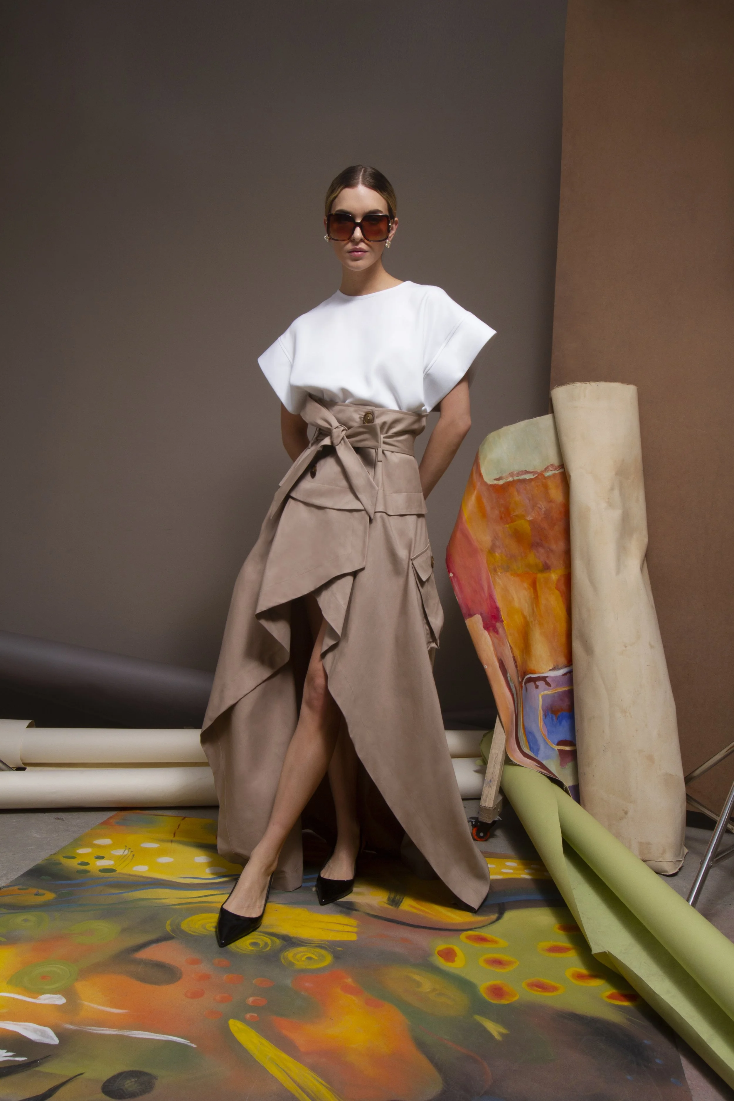 A woman wearing sunglasses, a white top, and a beige skirt poses in an art studio with large abstract paintings and colorful art supplies on the floor and leaning against the wall.