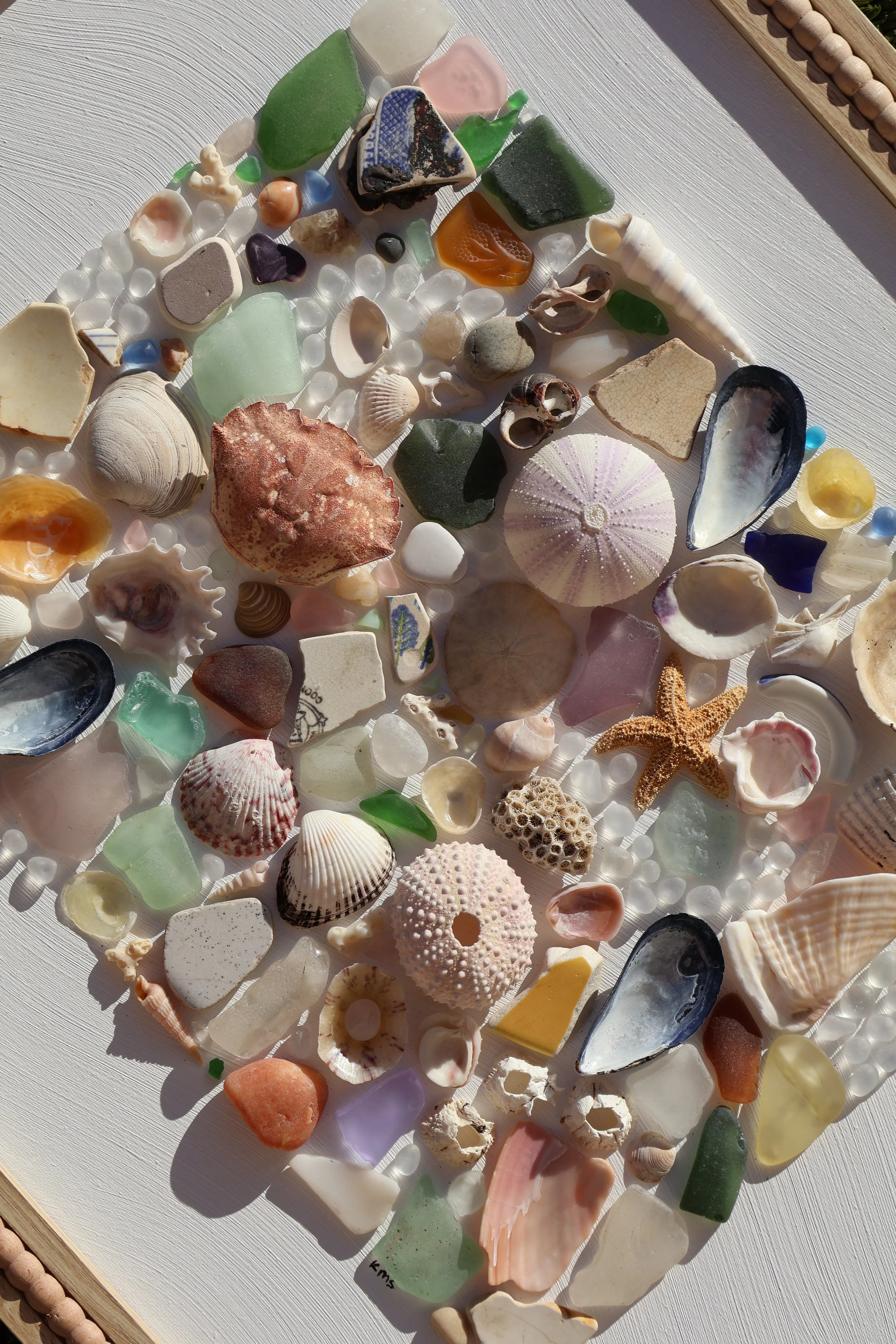 A collection of various seashells, sea glass, and small marine-shaped objects arranged on a white surface with a light wood frame.