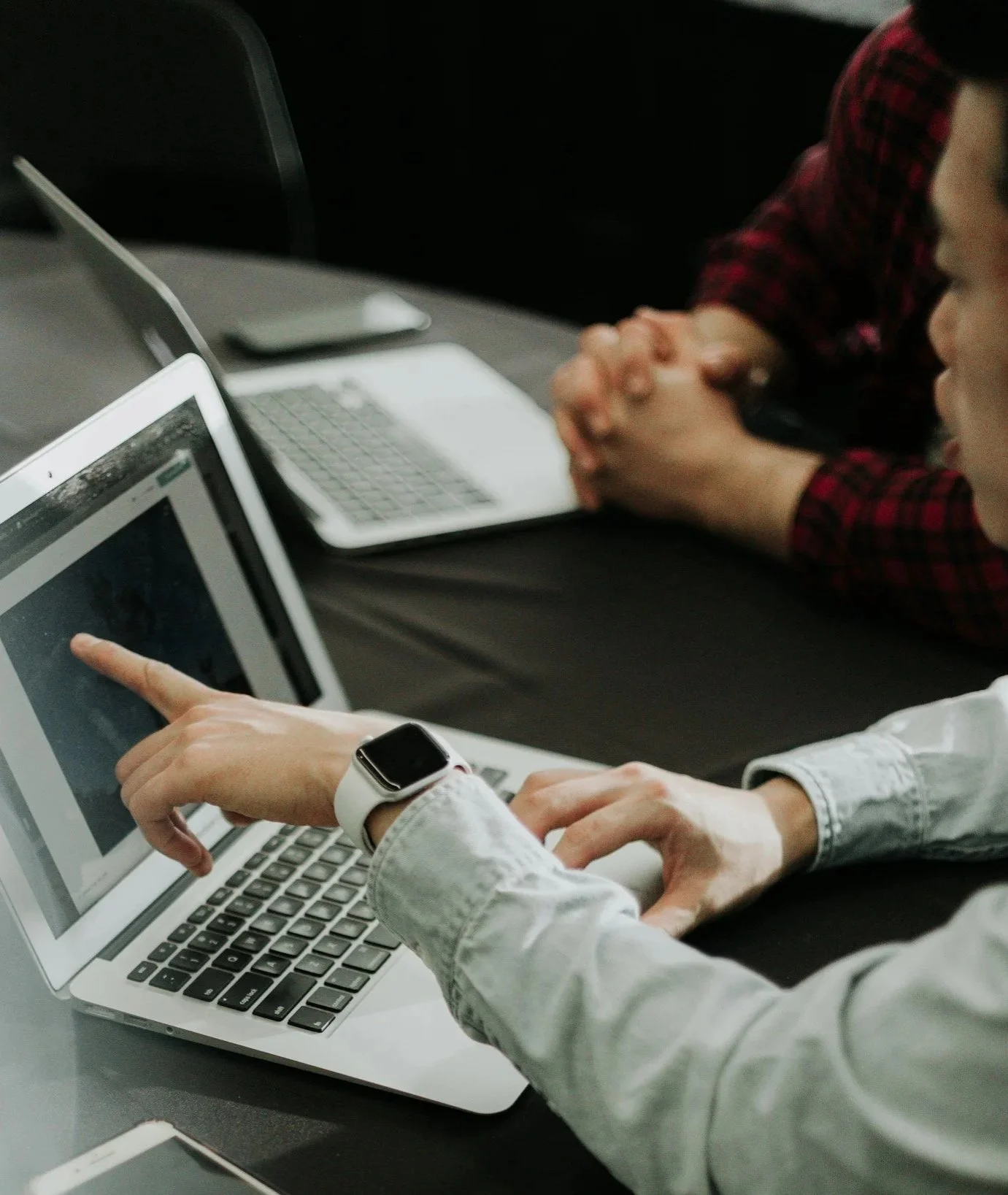 Person pointing at a laptop screen during a meeting, with other laptops and participants with clasped hands in the background.