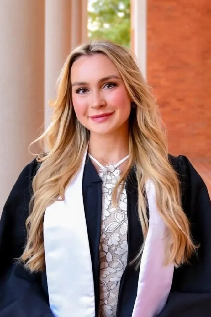 Young woman in graduation gown and stole, smiling with long blonde hair, standing indoors with a window and brick wall in the background.