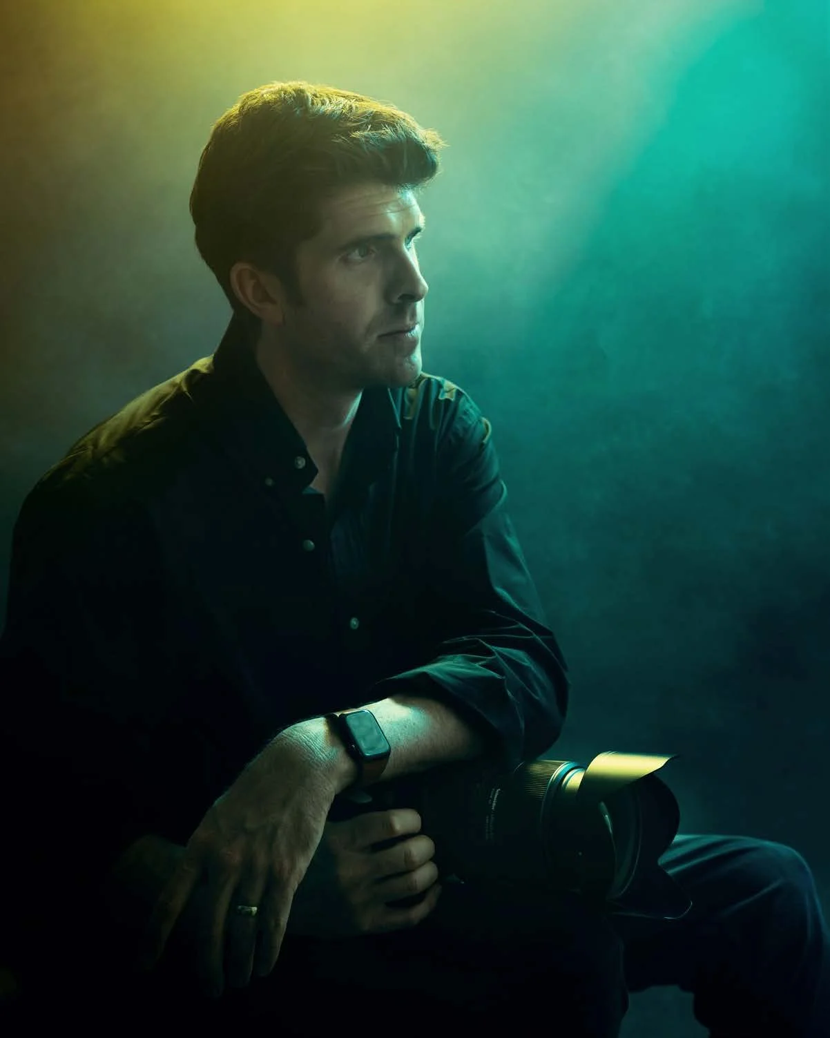 A man sitting in a dark setting with a gradient green and yellow background, wearing a black shirt and holding a camera, gazing thoughtfully to the side.