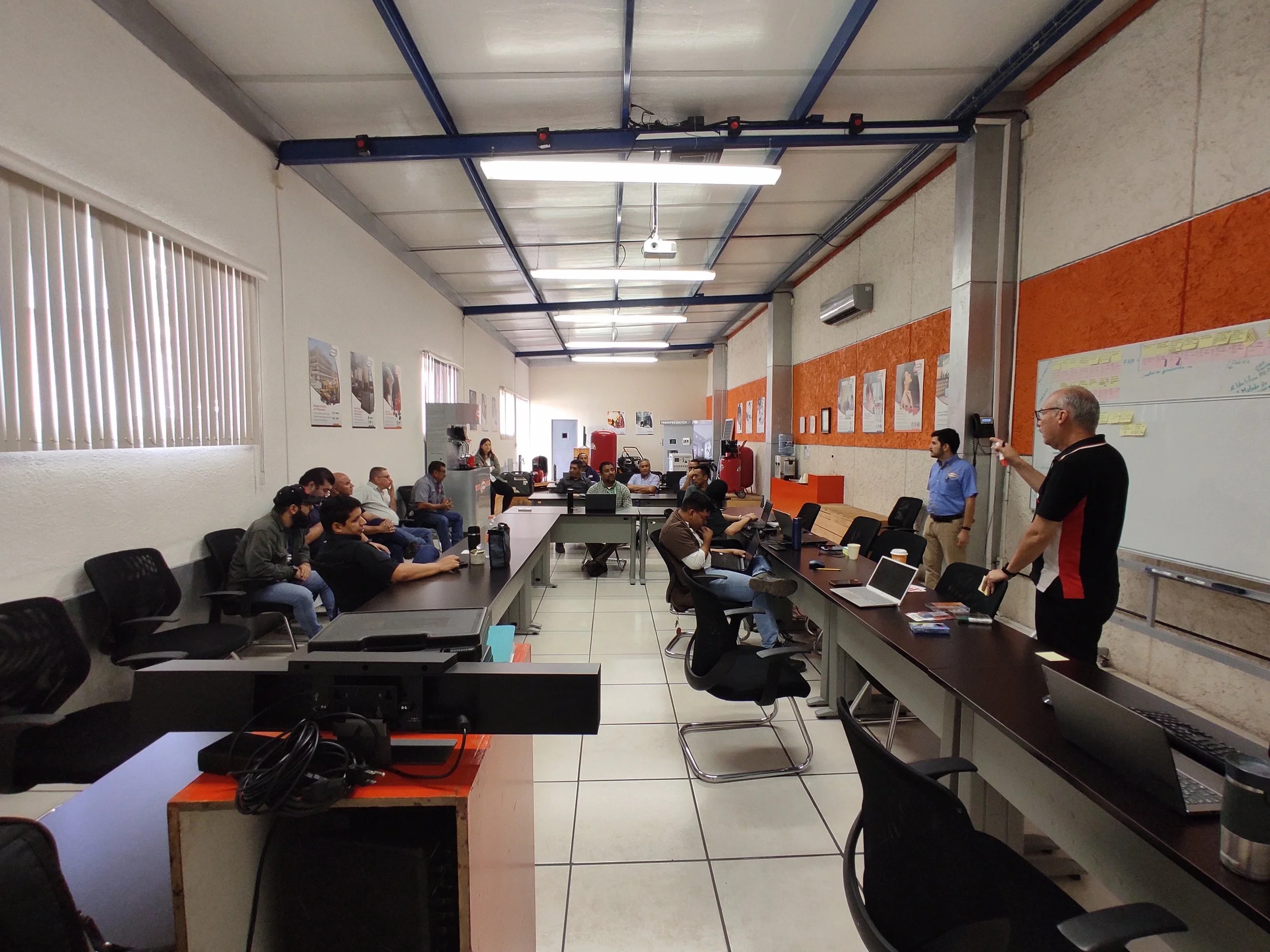 Sala de reuniones con varias personas sentadas, algunos con laptops abiertos, y una persona de pie hablando en frente, en una presentación. Hay pizarras blancas, posters en la pared y equipo de oficina.