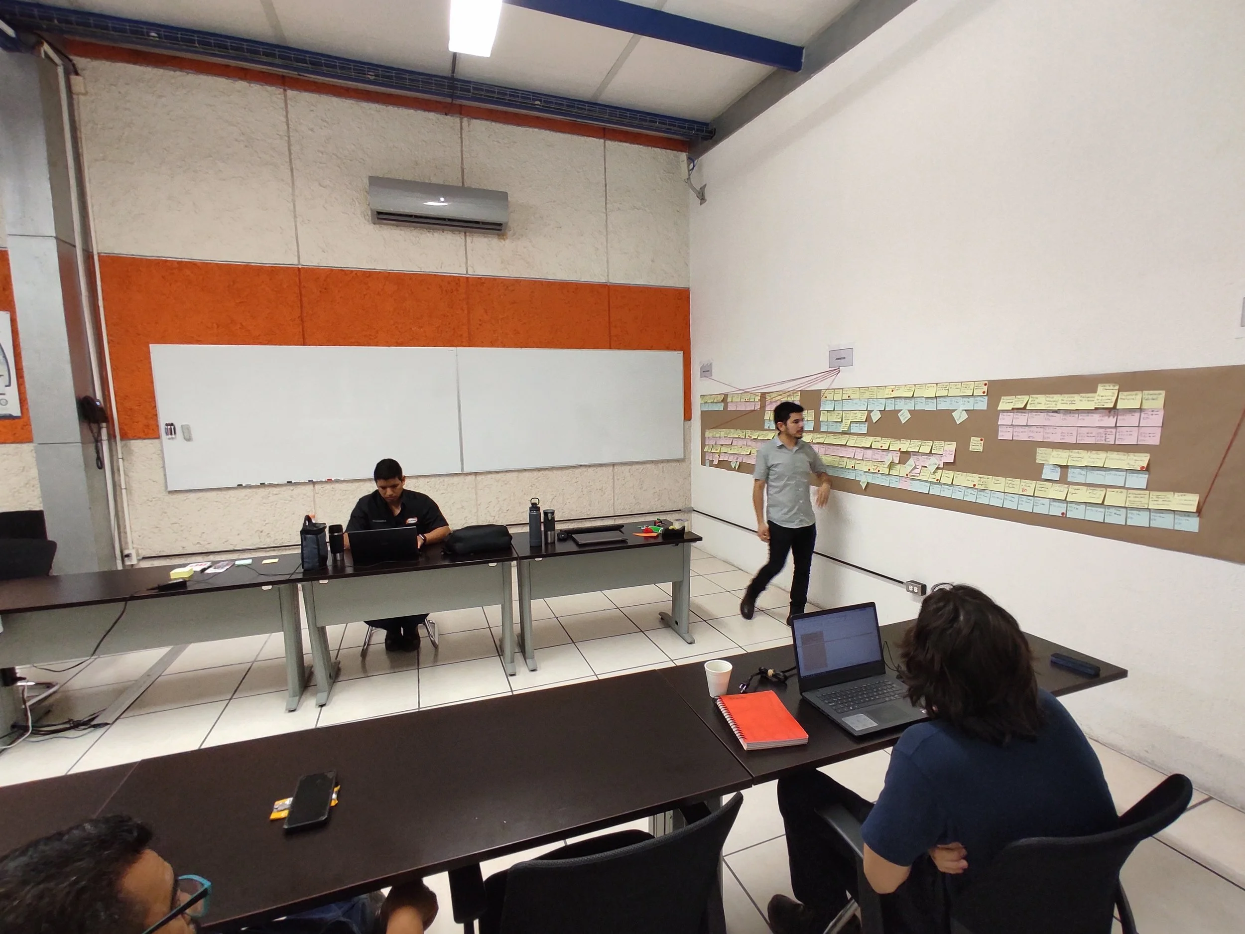 Un grupo de personas en una sala de reuniones participando en una presentación. Una persona está de pie frente a una pared con notas adhesivas, otra está sentada frente a un portátil, y otra más observa.