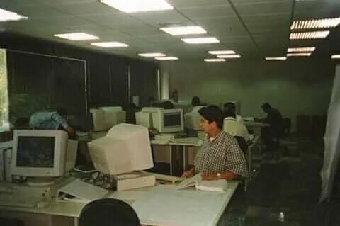 Una sala de computadoras de oficina con varias personas trabajando en computadoras antiguas CRT.