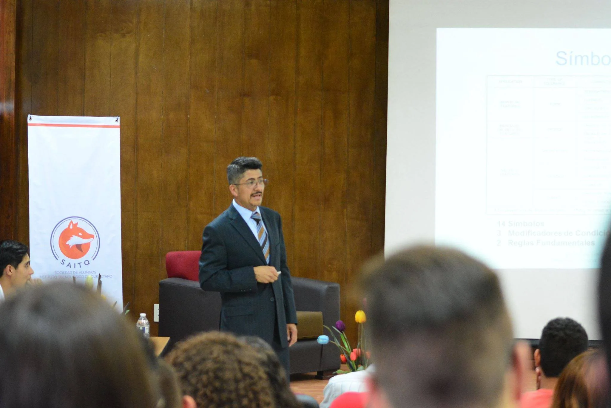Un hombre vestido con saco y corbata dando una presentación frente a una audiencia en un salón de clases o conferencias, con una pantalla y un cartel con el logotipo de SAITO en el fondo.