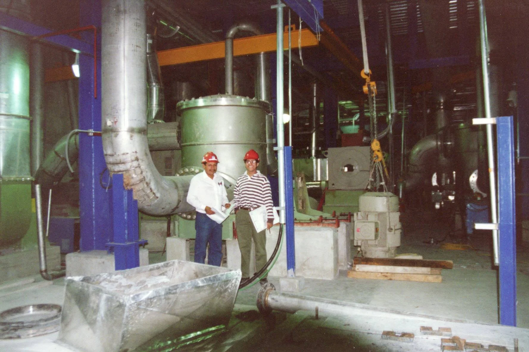 Dos hombres con cascos rojos de seguridad en una planta industrial, rodeados de maquinaria grande y tuberías.