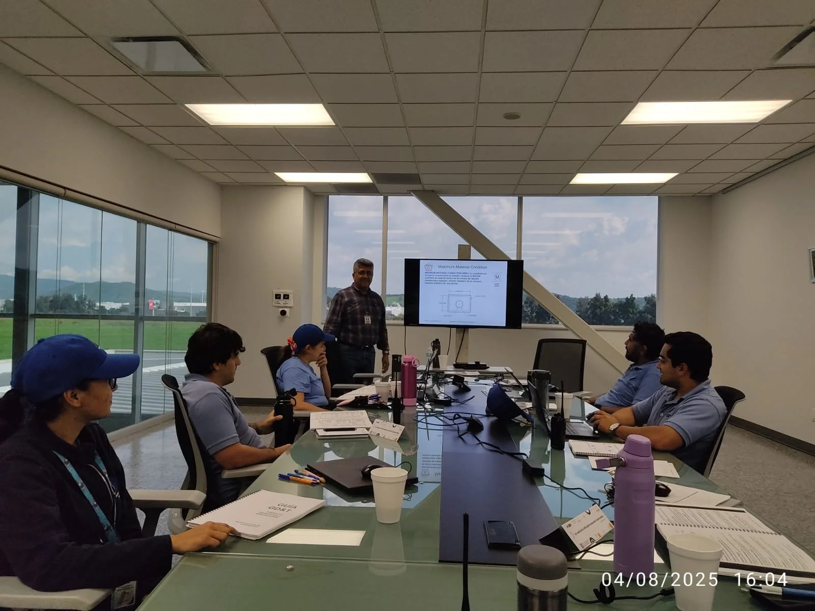 Reunión en una sala de conferencias con varias personas sentadas alrededor de una mesa y una persona de pie frente a una pantalla con una presentación. Ventanas grandes con vista al exterior.