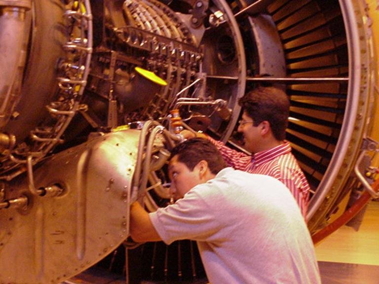Dos personas trabajando en una máquina grande, posiblemente una cámara de vacío o un equipo científico, en un entorno de laboratorio o museo técnico.