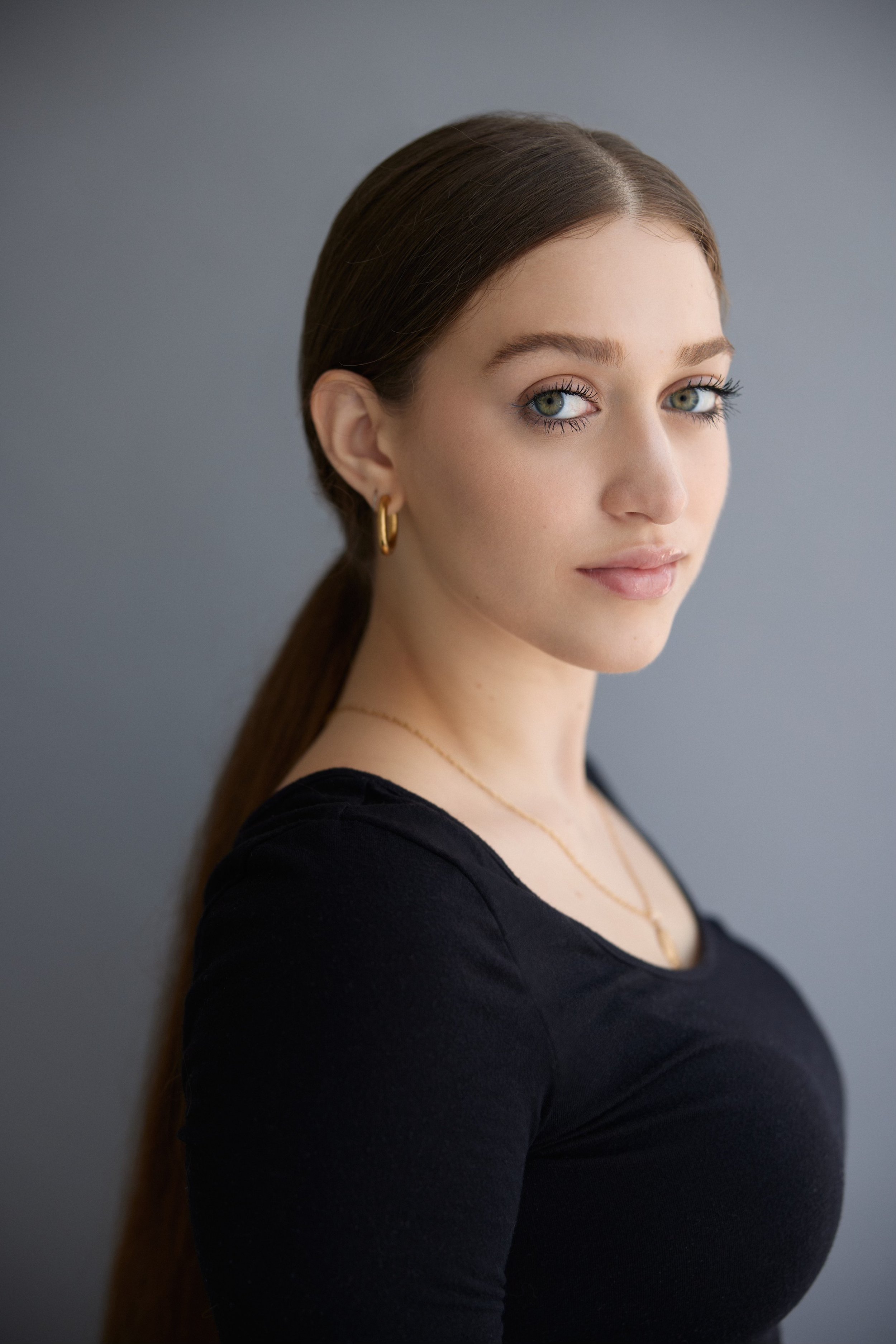 A young woman with long brown hair, wearing a black top and gold jewelry, standing against a plain gray background.