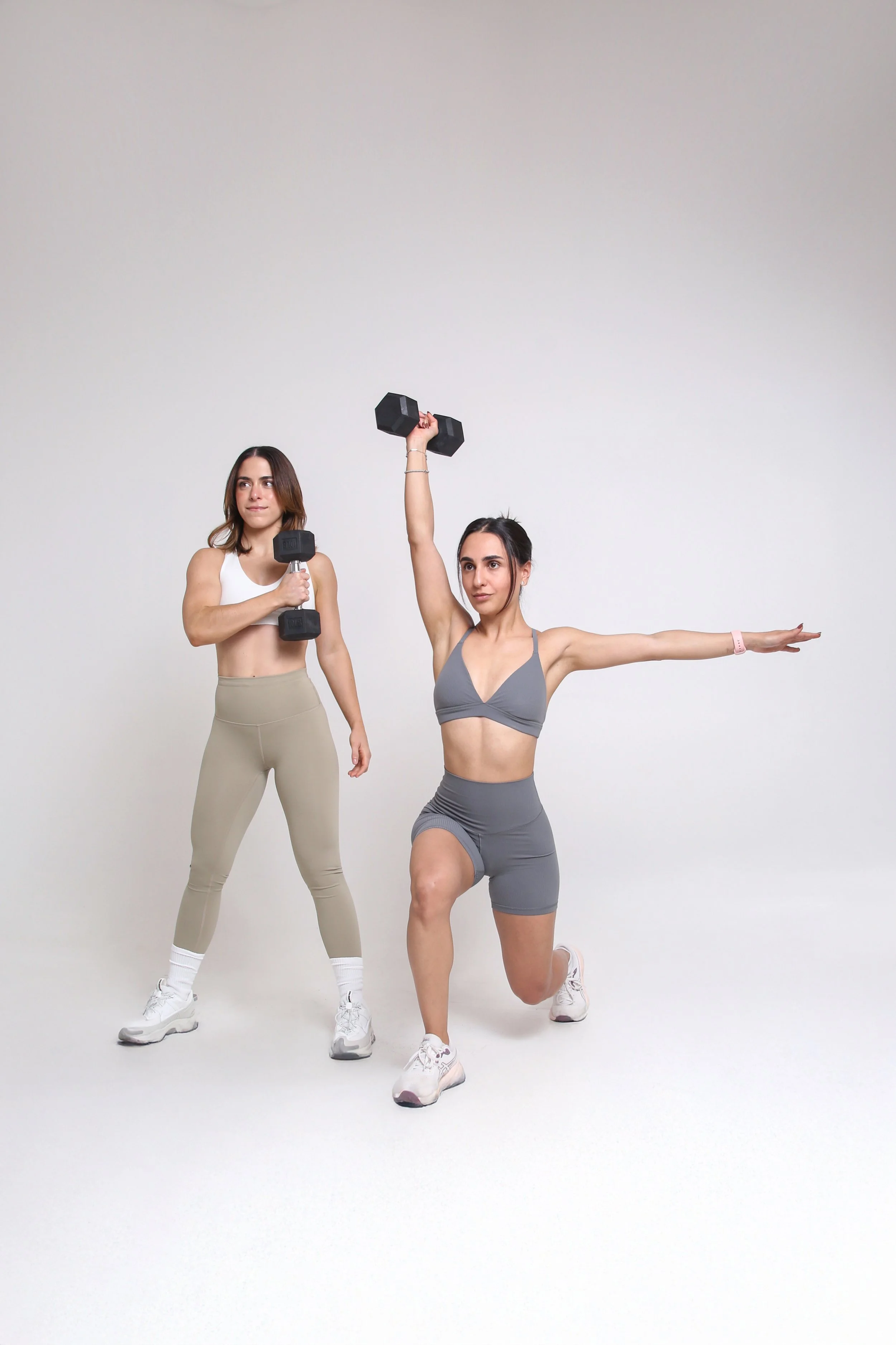 Dos mujeres haciendo ejercicio con pesas en un fondo blanco, una sosteniendo una pesa en cada mano y la otra levantando una pesa con una mano mientras está en una postura de yoga.