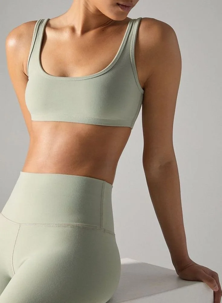 Mujer usando ropa deportiva de color beige, vista parcial del torso y las piernas, en un fondo gris claro.