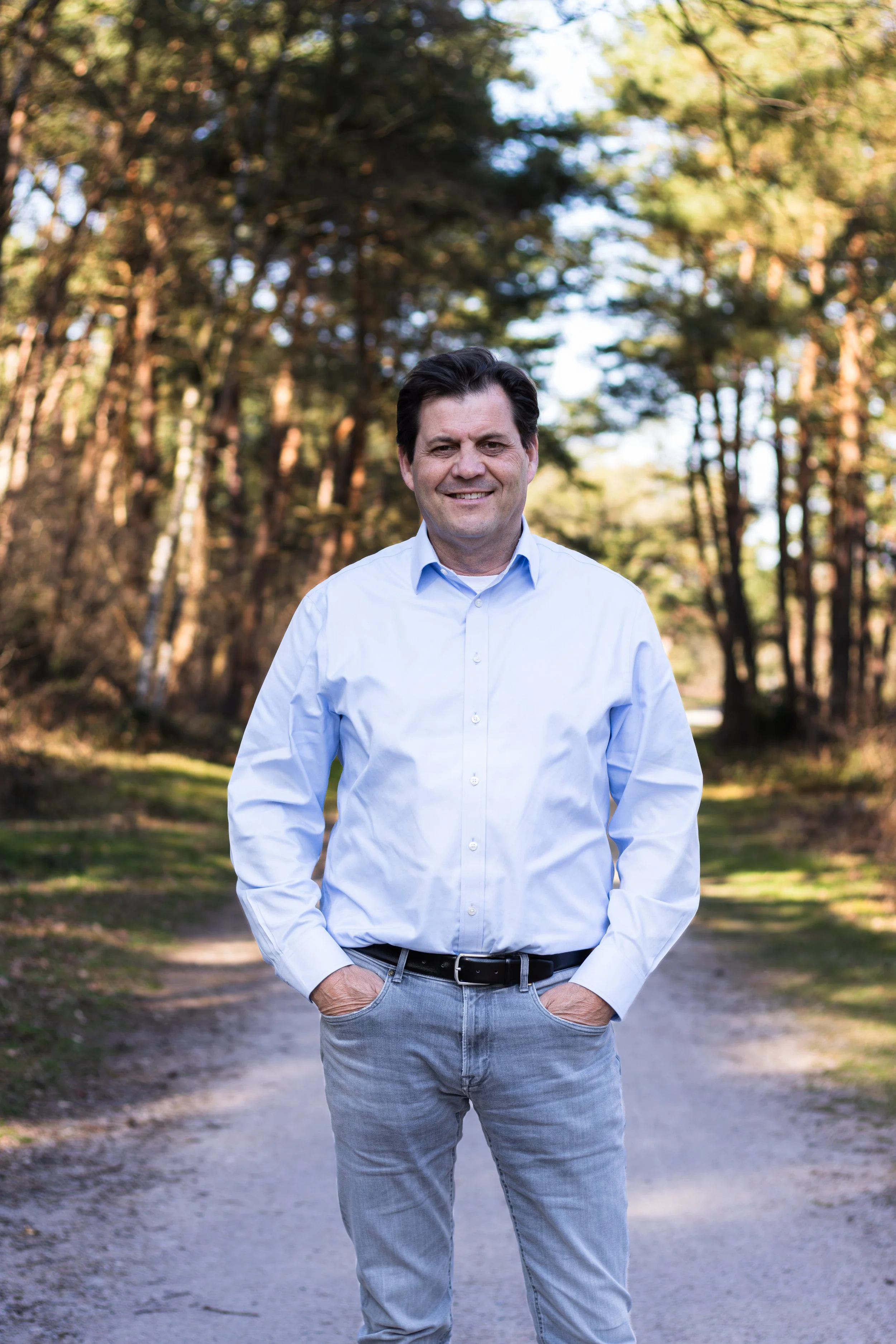 A man smiling outdoors in a park with trees in the background, wearing a light blue button-up shirt and gray pants