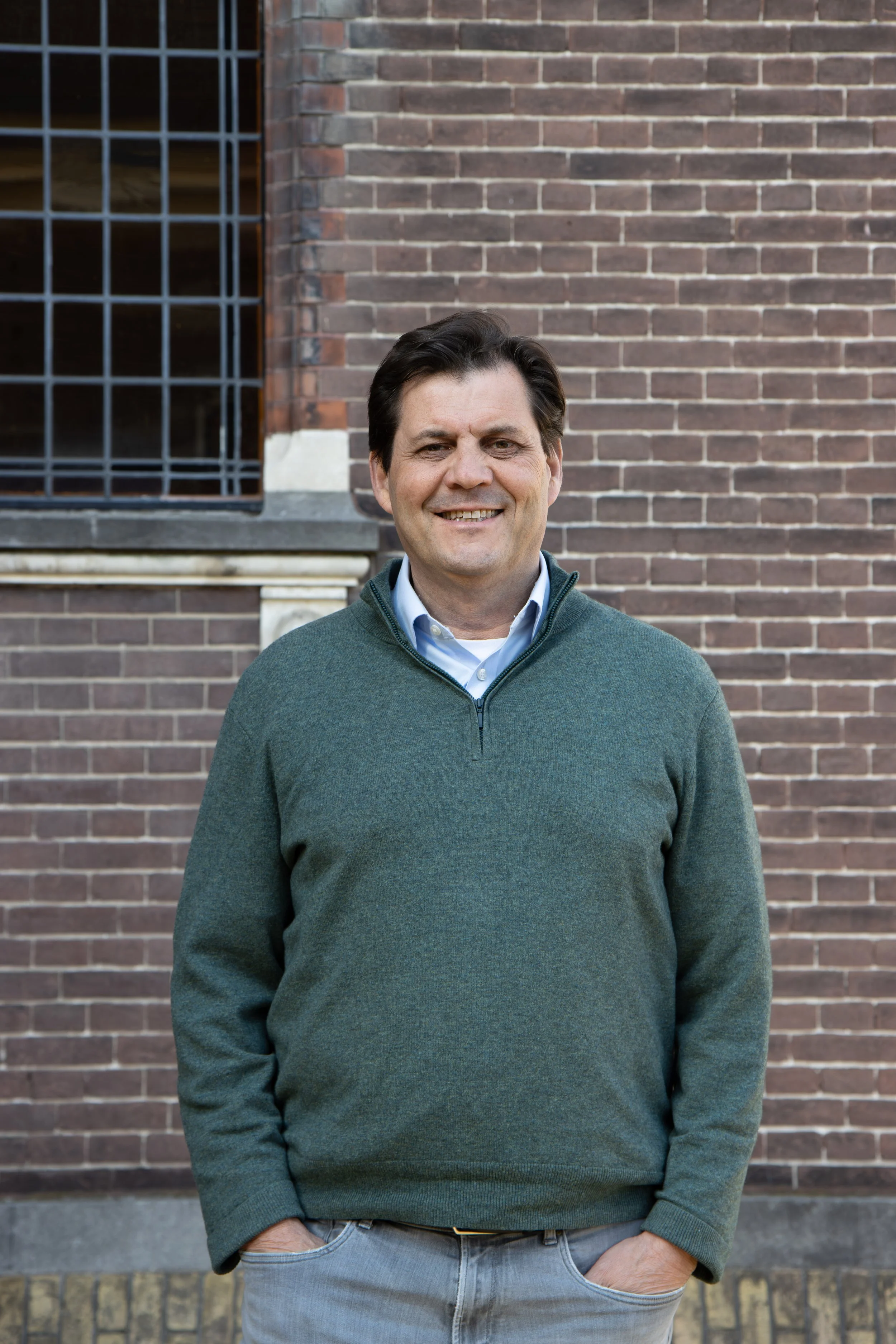 A man standing outdoors in front of a brick building, smiling, with one hand in his pocket and wearing a green sweater over a button-up shirt.