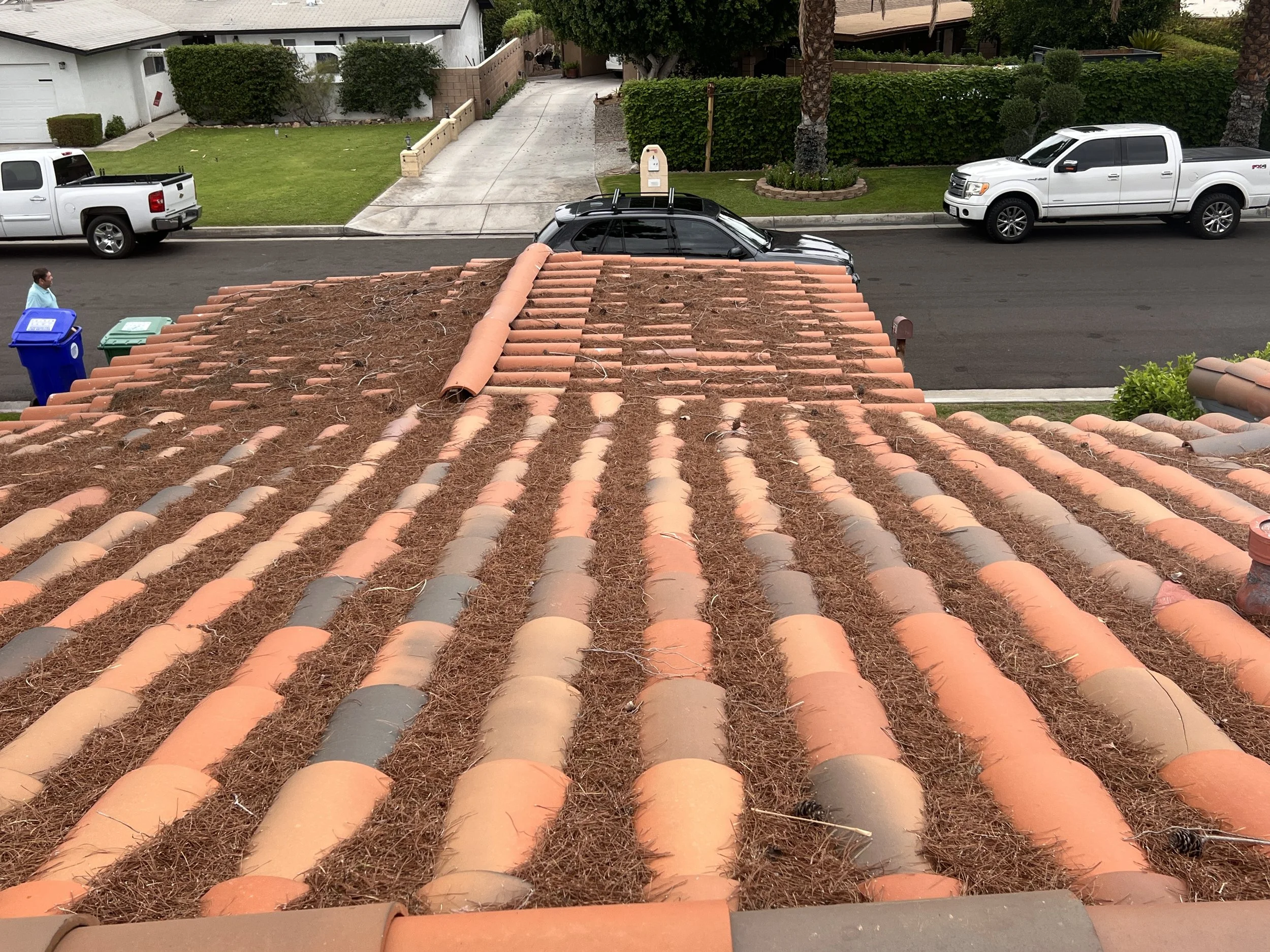 View of a rooftop with weathered red clay tiles; some tiles replaced or displaced, with dirt, debris, and pine needles on the roof. Street and parked cars are visible in the background.