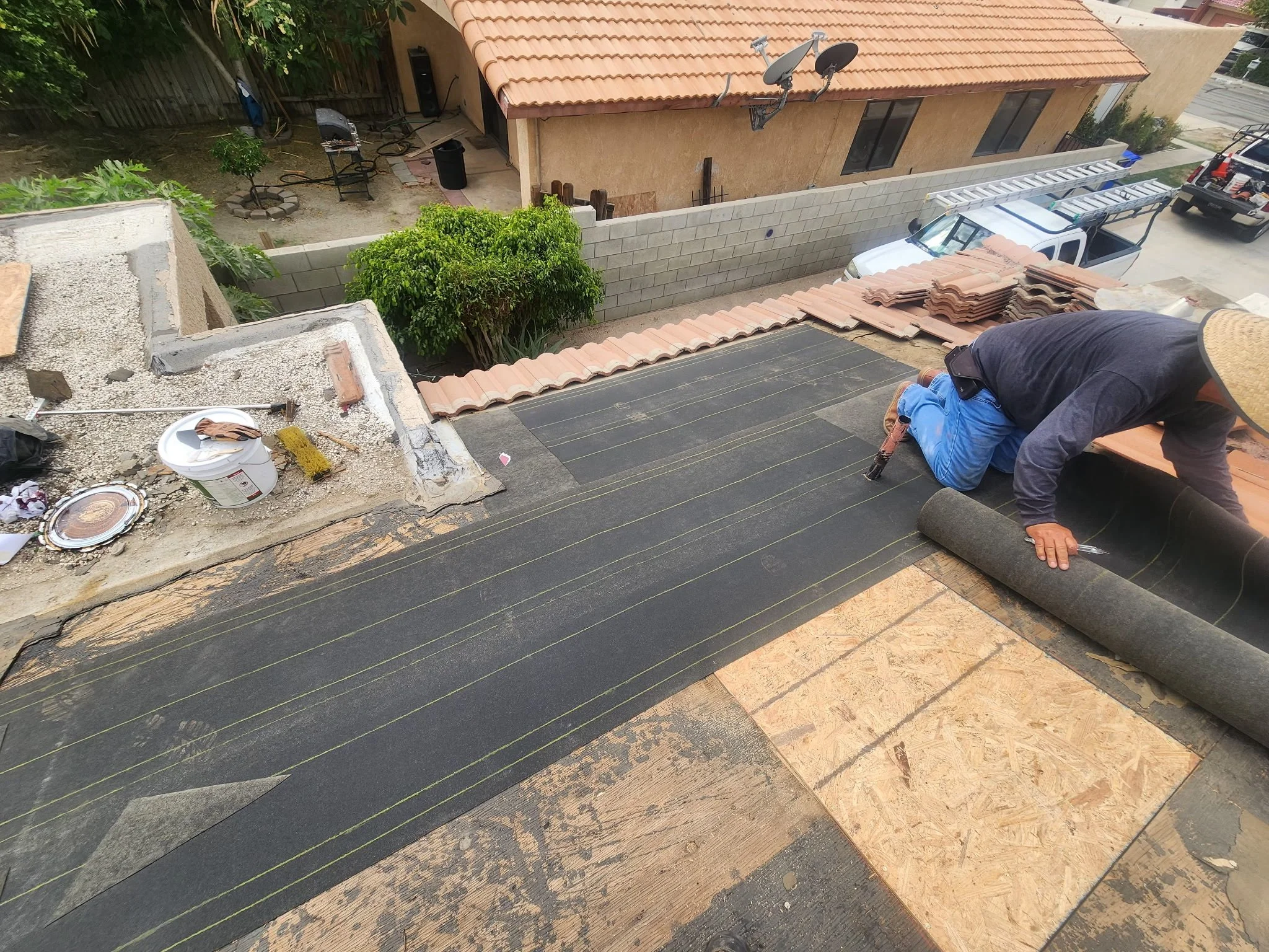 A worker installing roofing underlayment on a house roof, with laying tiles nearby, in an outdoor residential area.
