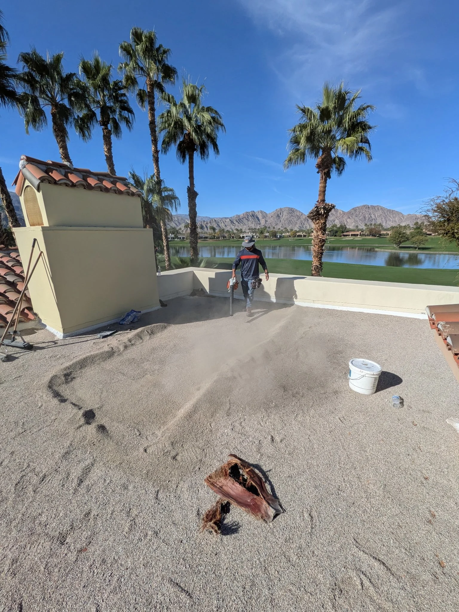 Construction worker on a rooftop with palm trees, mountains, and a lake in the background. Debris and a bucket are visible on the roof.
