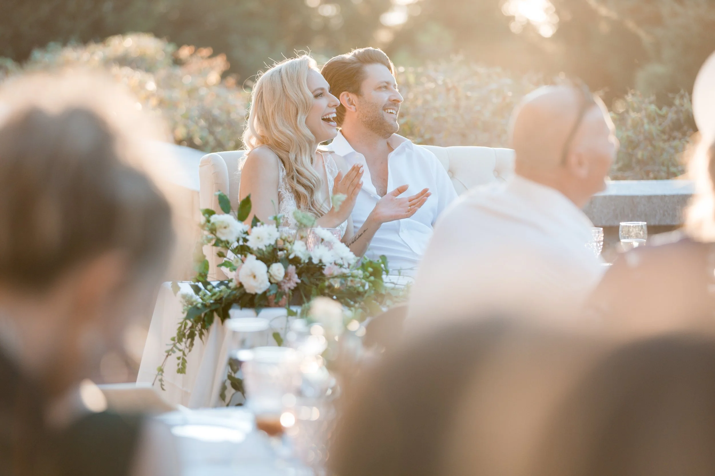 A couple laughing and smiling at an outdoor wedding reception during sunset, sitting behind a floral centerpiece with guests around, in warm light.
