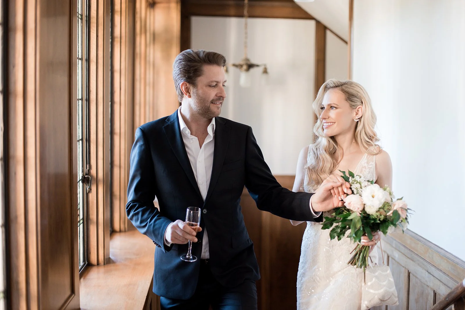 A man in a dark suit and a woman in a white wedding dress smiling and holding hands, with the woman holding a bouquet of flowers, standing inside a wooden-paneled room with large windows.