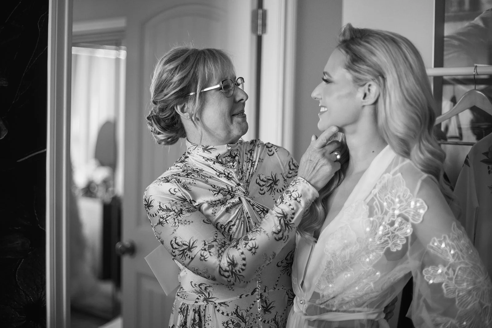 A mother is adjusting a necklace on her daughter the bride to be who is smiling, with her chin slightly tilted up. The scene appears intimate, pre-wedding preparation, in a dressing area in Vancouver taken by Allison Taylor Creative
