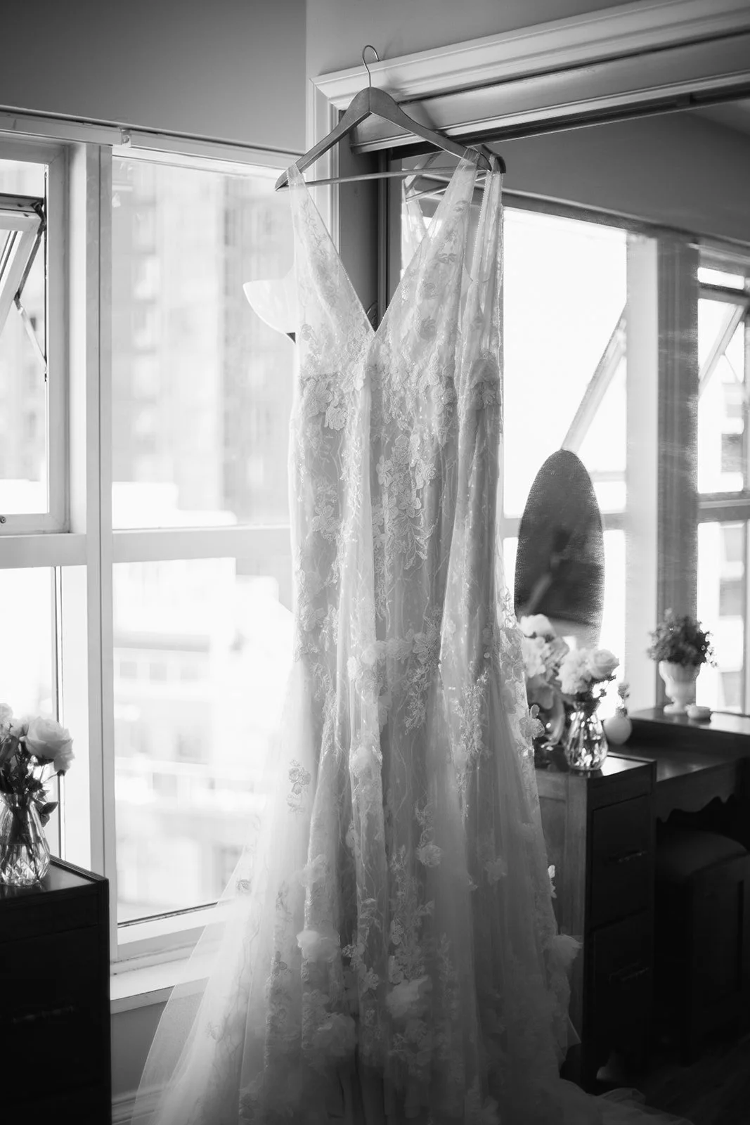 A lace wedding dress hanging on a hanger against a window with sunlight streaming in, with flowers in vases on nearby furniture before the wedding at UBC botanical garden