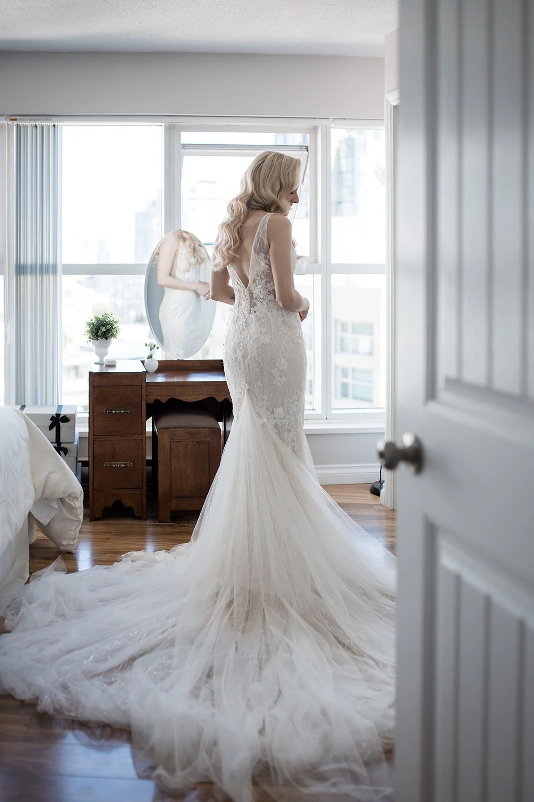 A bride in a lace wedding dress with a long train standing in front of a mirror in a bright room shot by Allison Taylor Creative