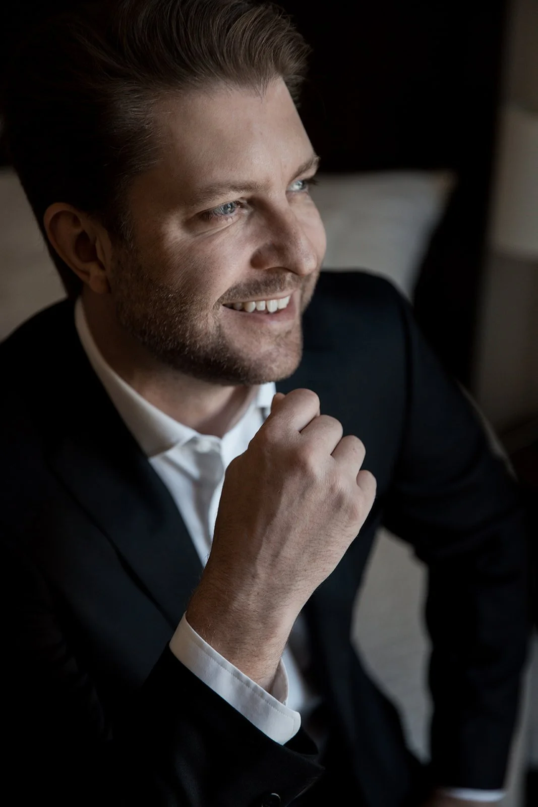 A smiling groom with blue eyes, light brown hair, and a beard, dressed in a black suit and white shirt, sitting indoors and looking out of a window.