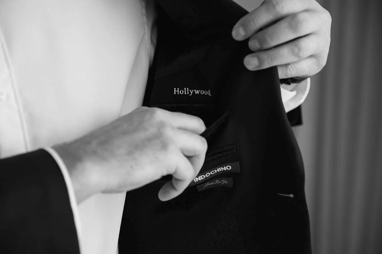 Close-up of a groom in a tuxedo, adjusting their tuxedo jacket which has a label that reads "Hollywood."