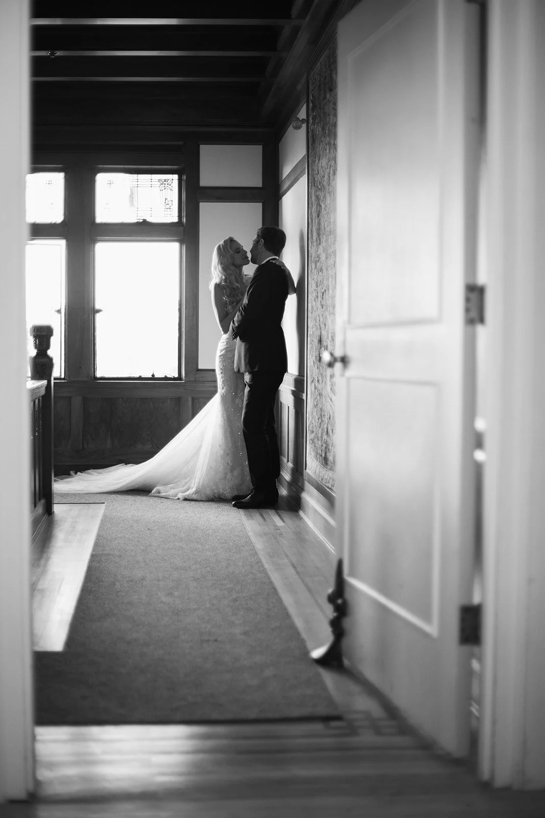 A black and white photo of a bride and groom standing close together inside a house, facing each other, near a window, with the bride in a wedding gown and the groom in a suit.