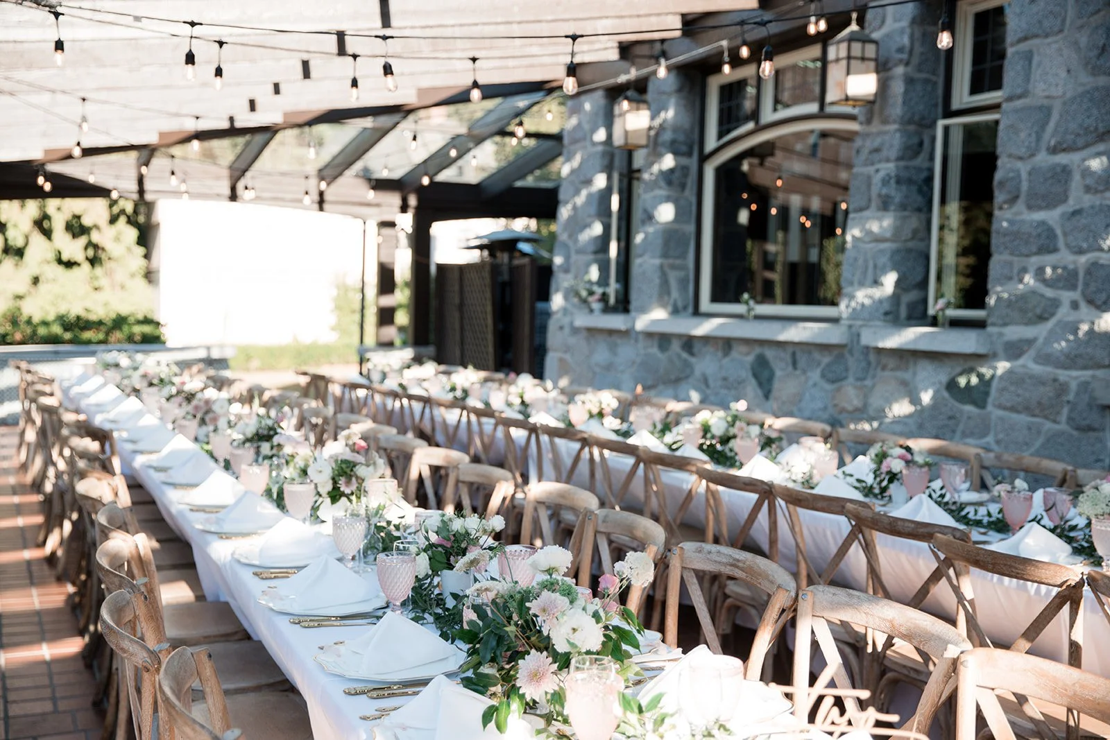 Long outdoor dining table set with white tablecloths, pink and white floral arrangements, white napkins, glasses, and gold utensils under string lights, beside a stone building.