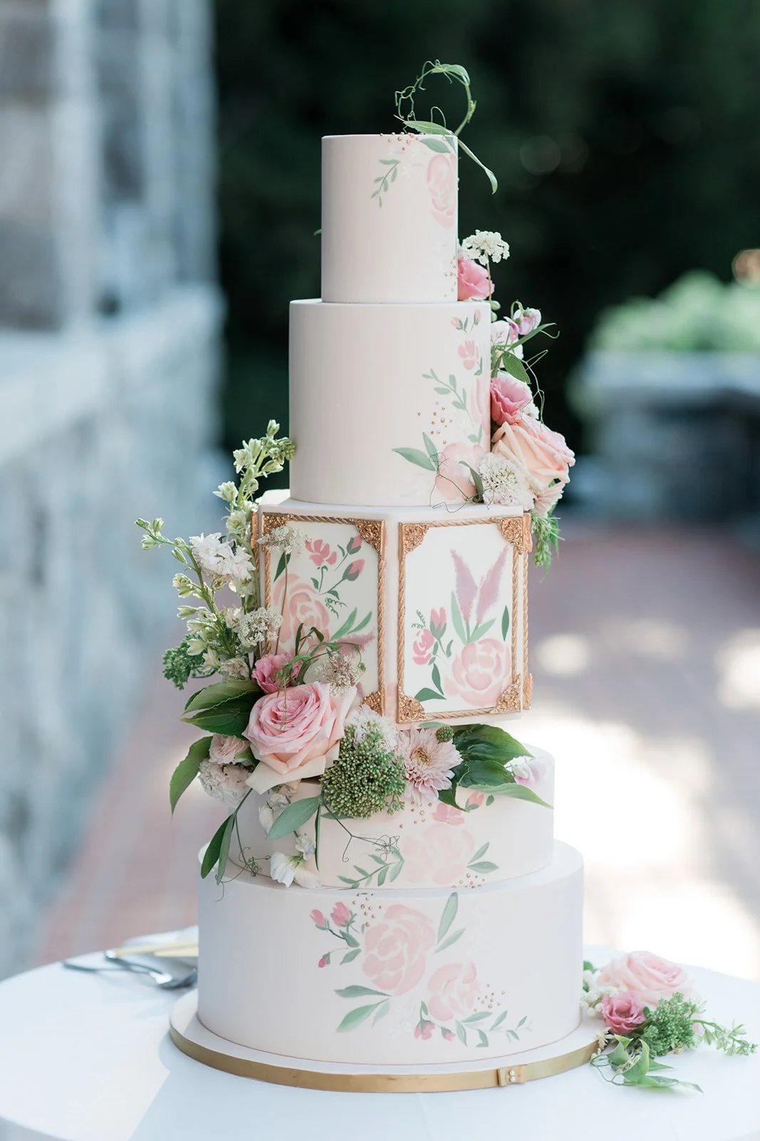 A four-tier wedding cake decorated with pink and white floral patterns, gold accents, and fresh flowers including pink roses and greenery.