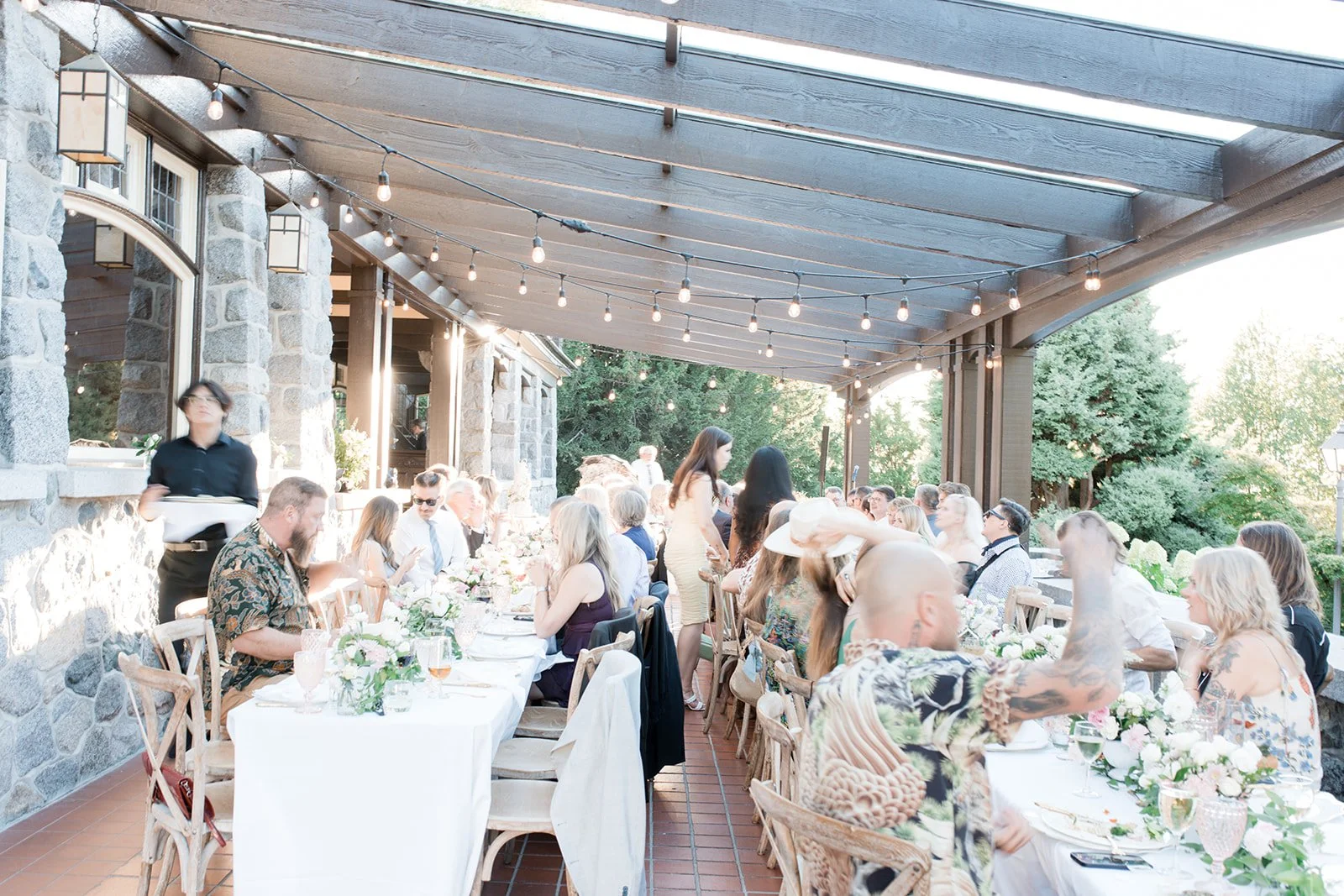 People gathered on a covered outdoor patio dining at a wedding or celebration with string lights and floral centerpieces.