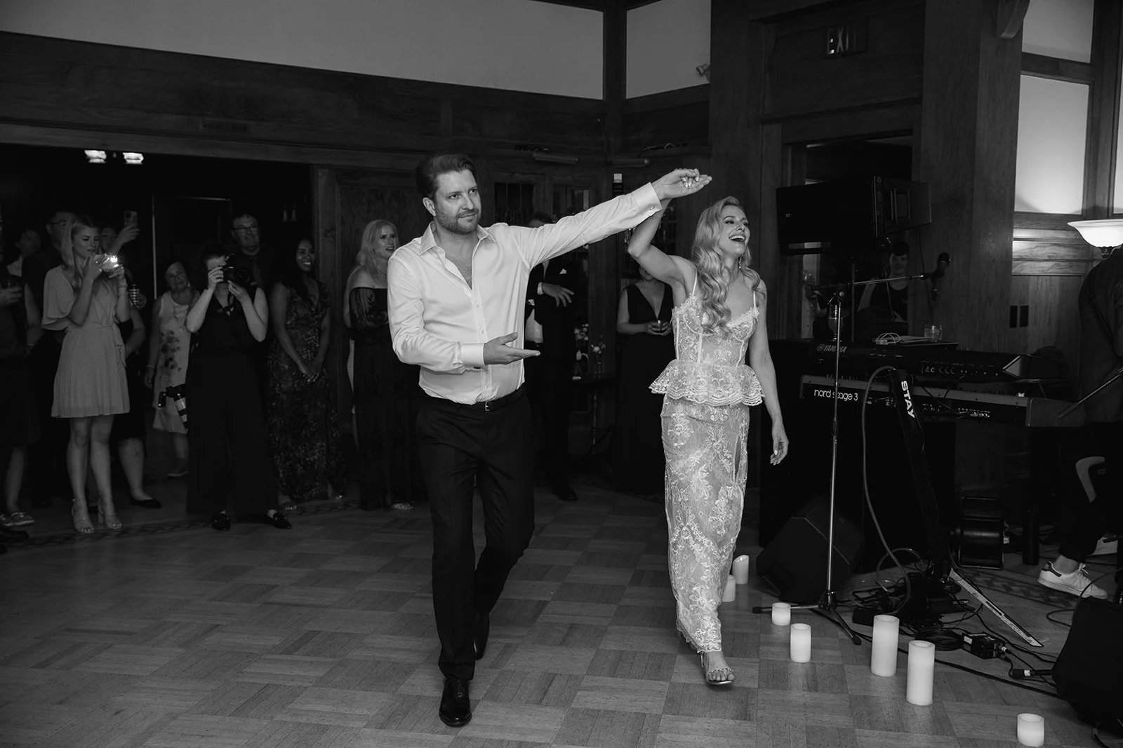 A man and woman dancing at a wedding reception, surrounded by guests, with candles on the floor and musical equipment nearby.