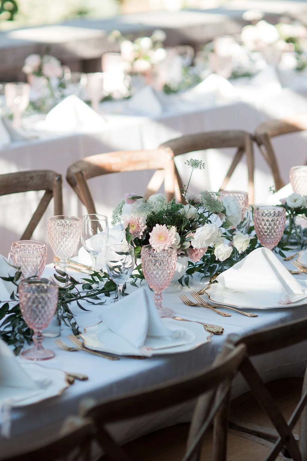 Elegant outdoor dining table decorated with white napkins, pink and clear glassware, gold cutlery, and a floral centerpiece of white and pink flowers with greenery, set for a special event or celebration.