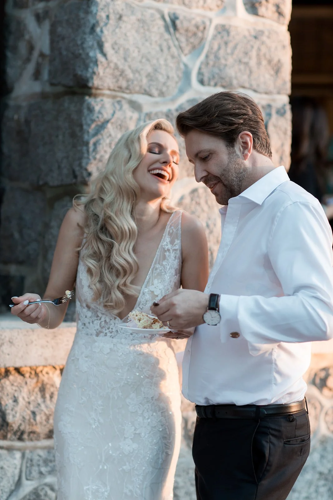 A bride in a white wedding dress and a groom in a white shirt are smiling and sharing a piece of cake during a wedding celebration outdoors, near a stone wall.
