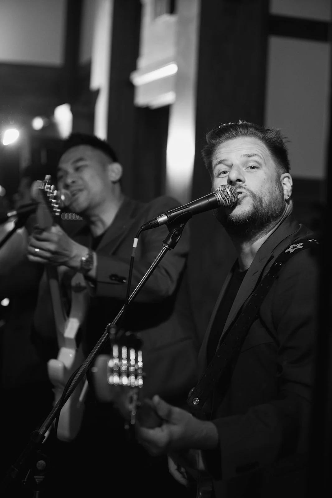 Black and white photo of two male musicians performing with one singing into a microphone and the other playing an acoustic guitar.
