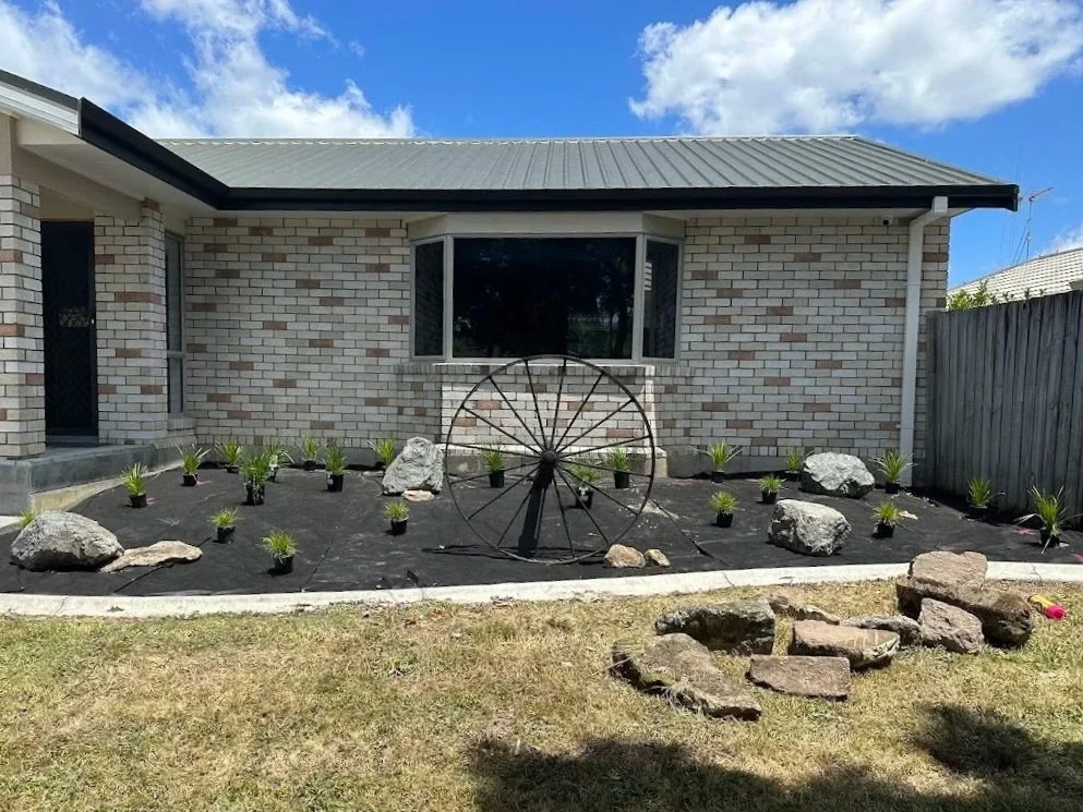Front yard with a brick house, newly landscaped garden, black mulch, small plants in pots, large decorative rocks, an old wheel sculpture, and a stone border with grass.