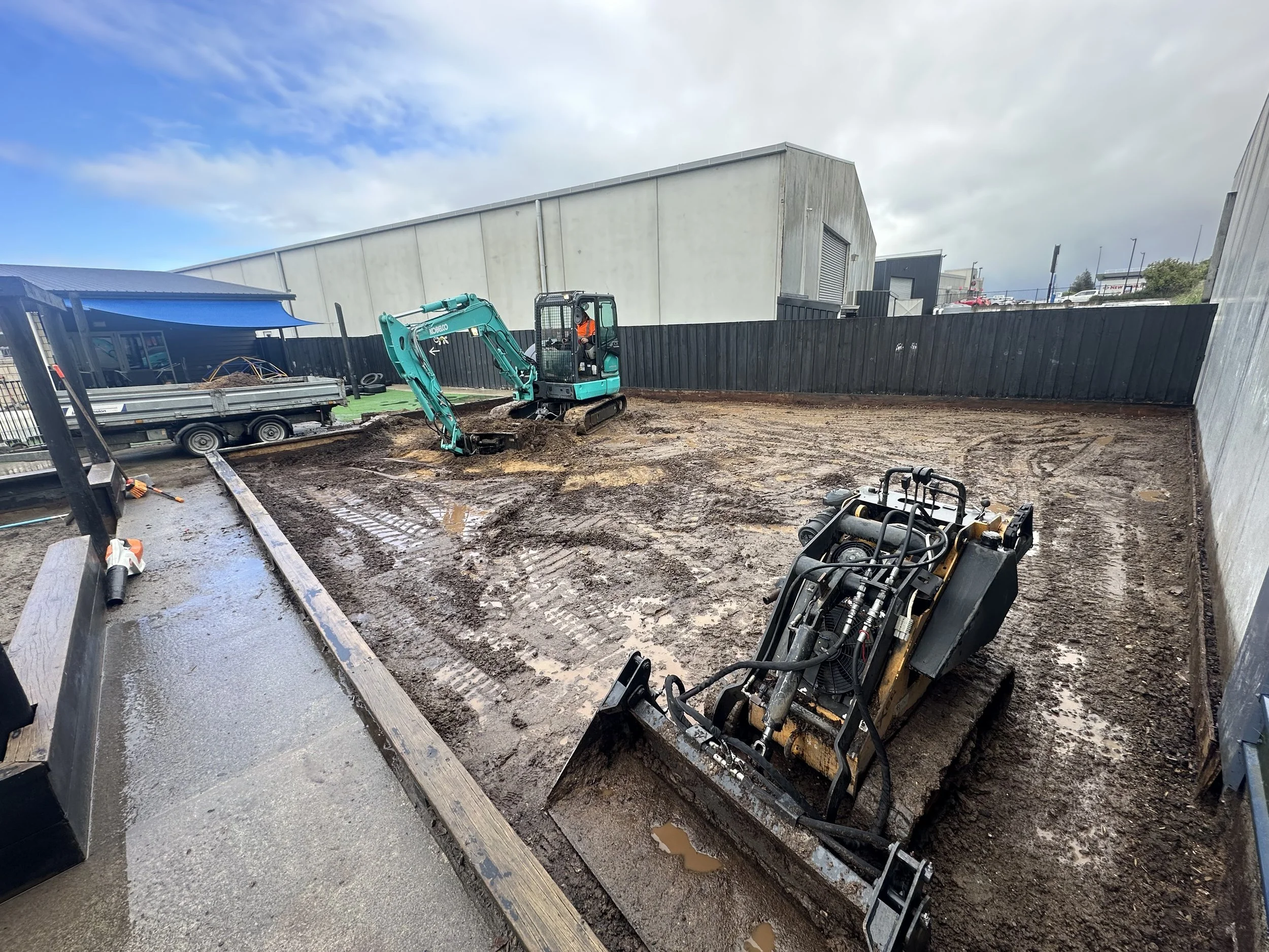 Construction site with dirt and excavation machinery, fenced perimeter, and industrial buildings in the background.