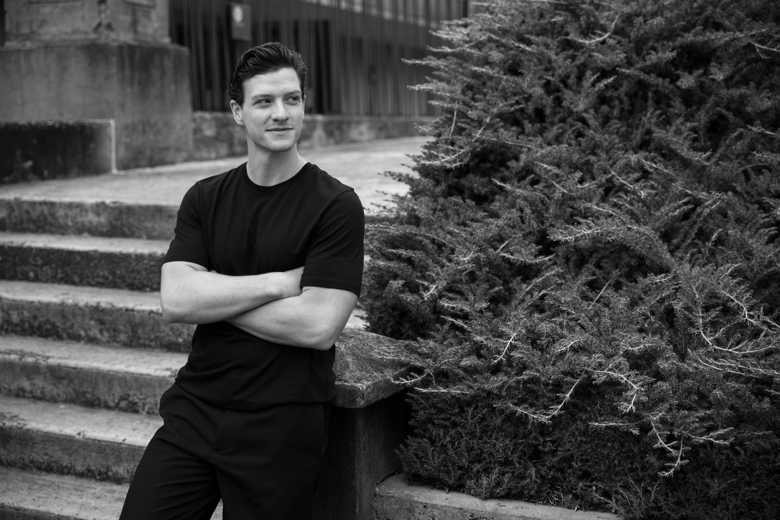 Atelier G Founder Will Gallagher in Black and White, arms crossed ligth smile in front of a pine and staircase