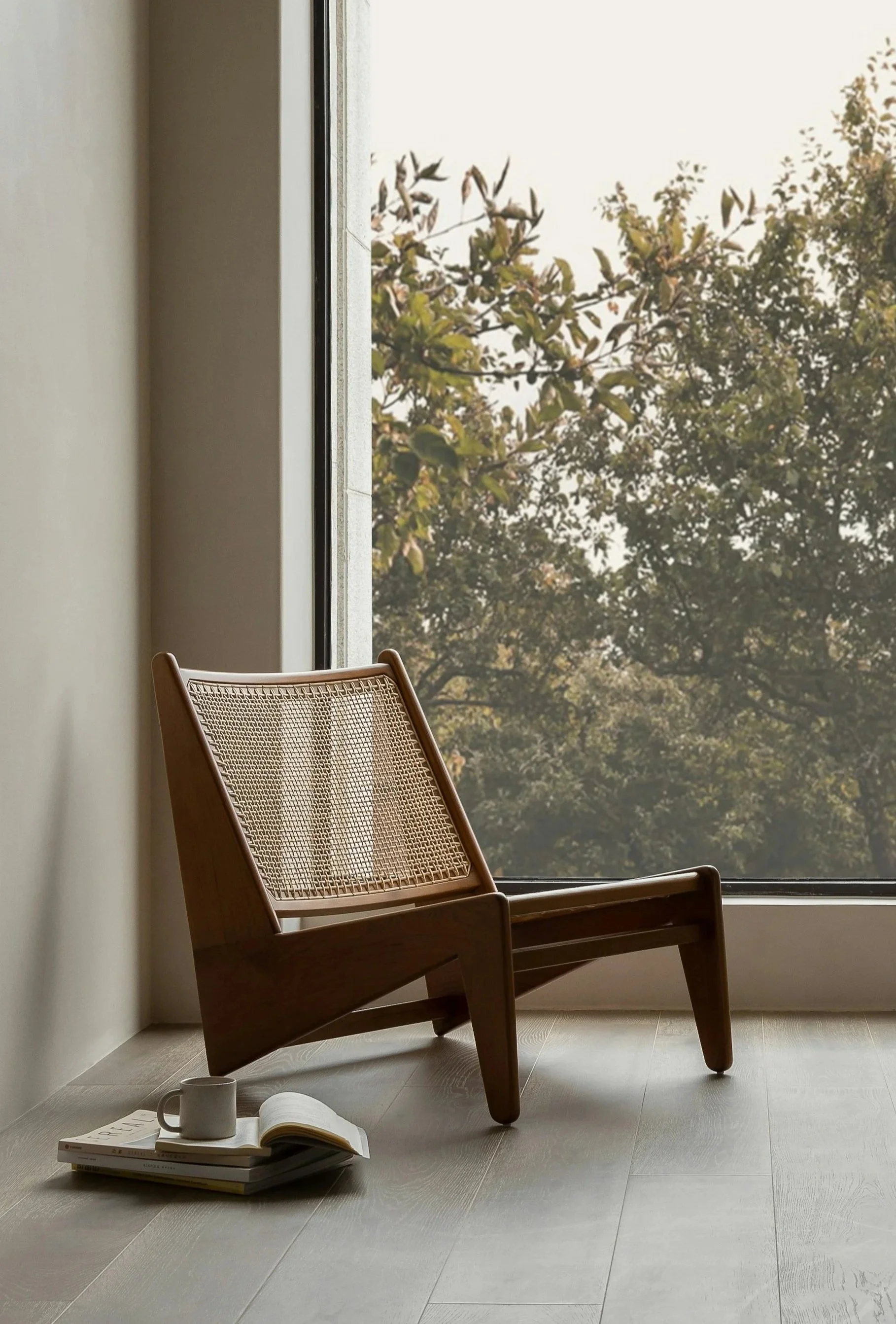 A wooden chair with a woven cane backrest by a large window showing trees outside, with a stack of books, a mug, and an open book on the floor.