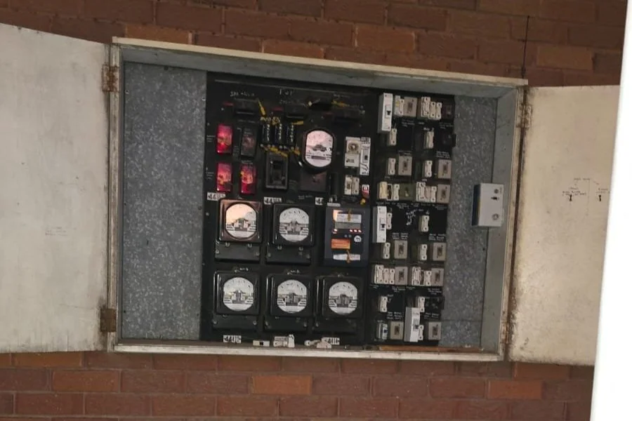 Electrical meters and circuit breakers inside an electrical panel box mounted on a brick wall.