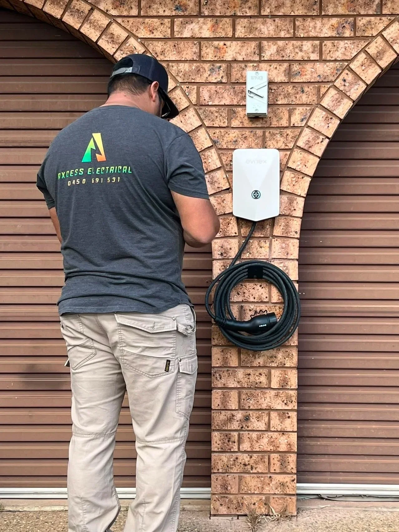 A man working on framing in a building under construction, wearing a gray t-shirt with a company logo and phone number, standing near a window.