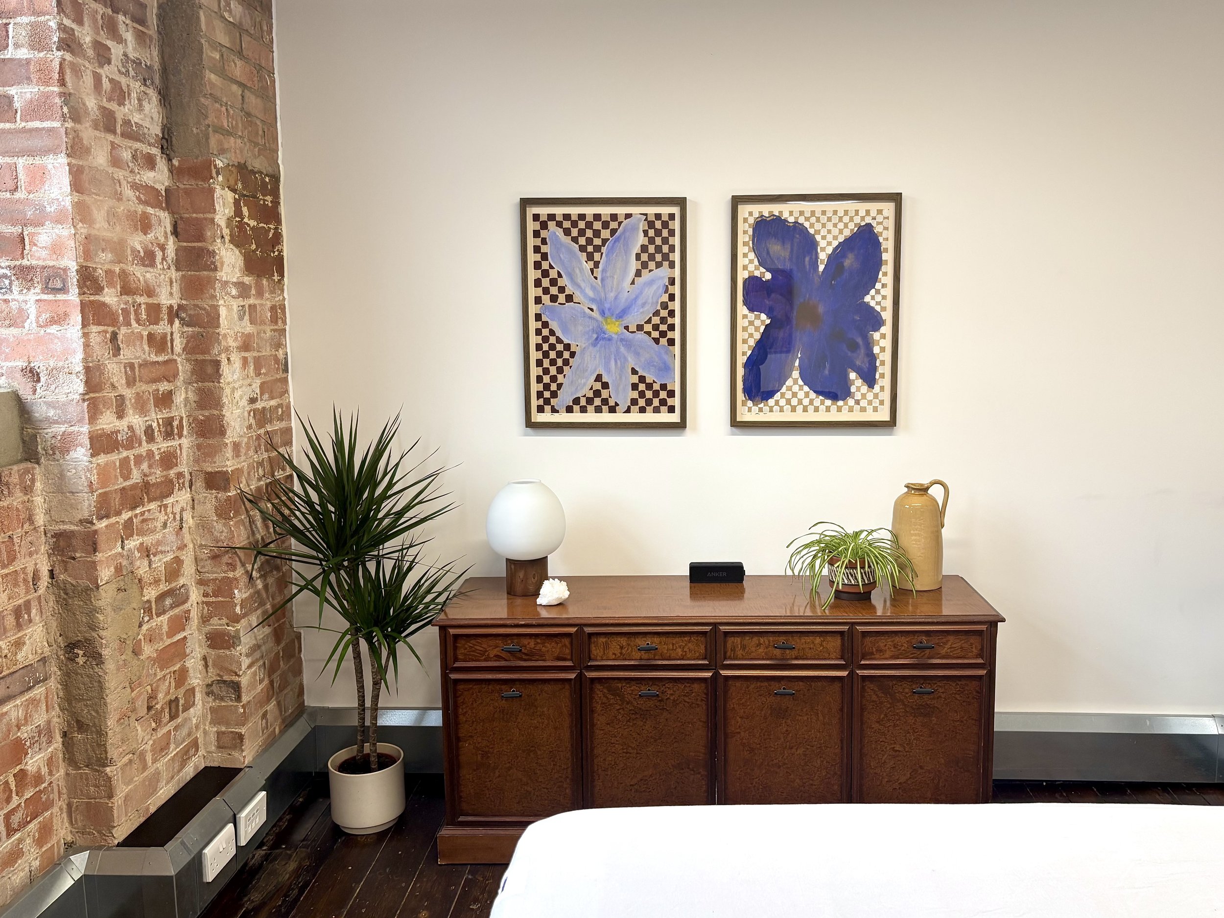 A wooden sideboard with a white table lamp, potted plant, a yellow pitcher, and a black wireless device on top. Two framed floral art prints hang above. There is a brick wall on the left and a plain beige wall. A houseplant in a white pot is on the floor to the left of the sideboard.
