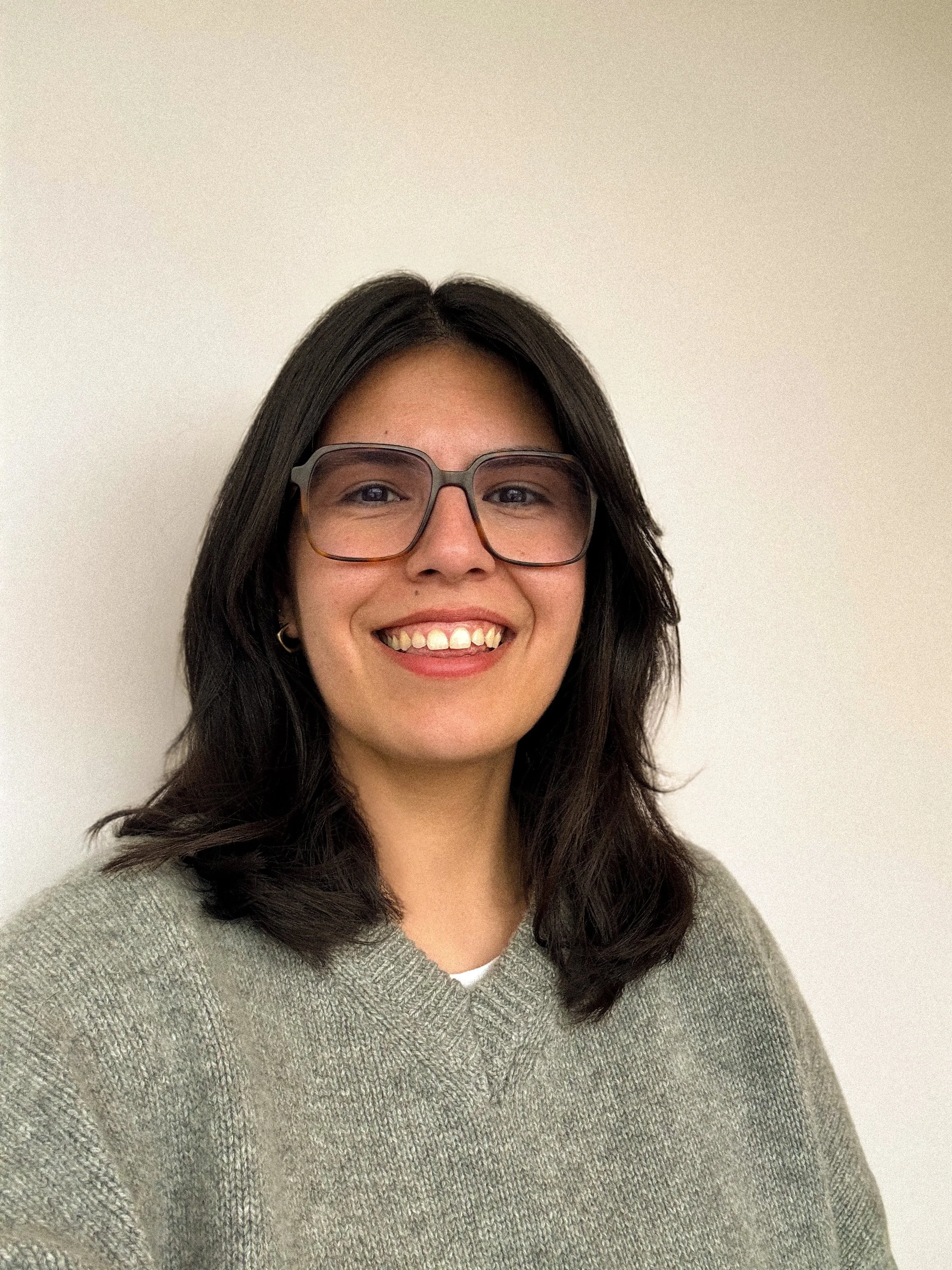 A woman with dark hair, glasses, and a gray sweater smiling in front of a plain light-colored wall.