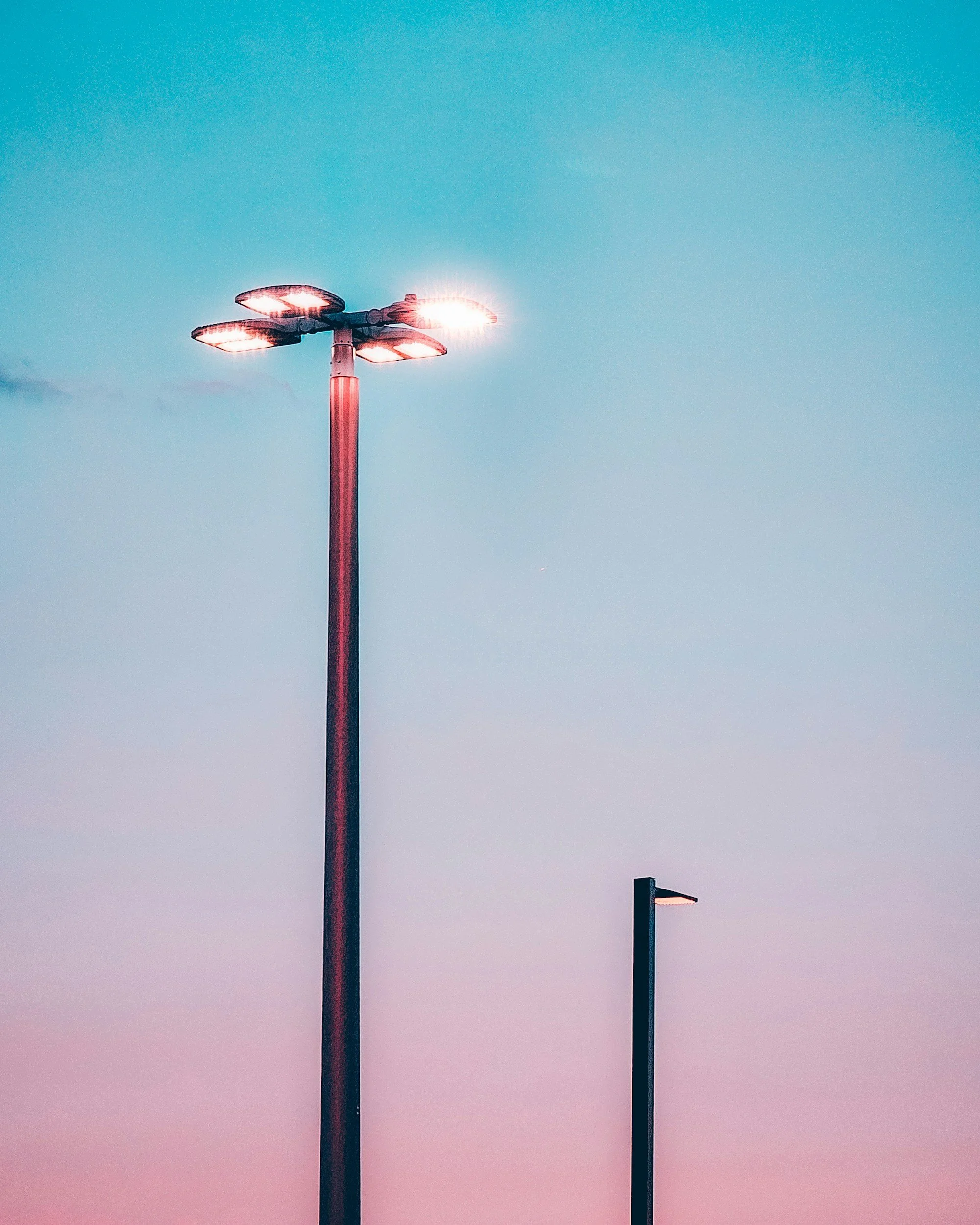A tall streetlight with multiple lamps at the top is illuminated against a colorful sky, accompanied by a shorter streetlight nearby.