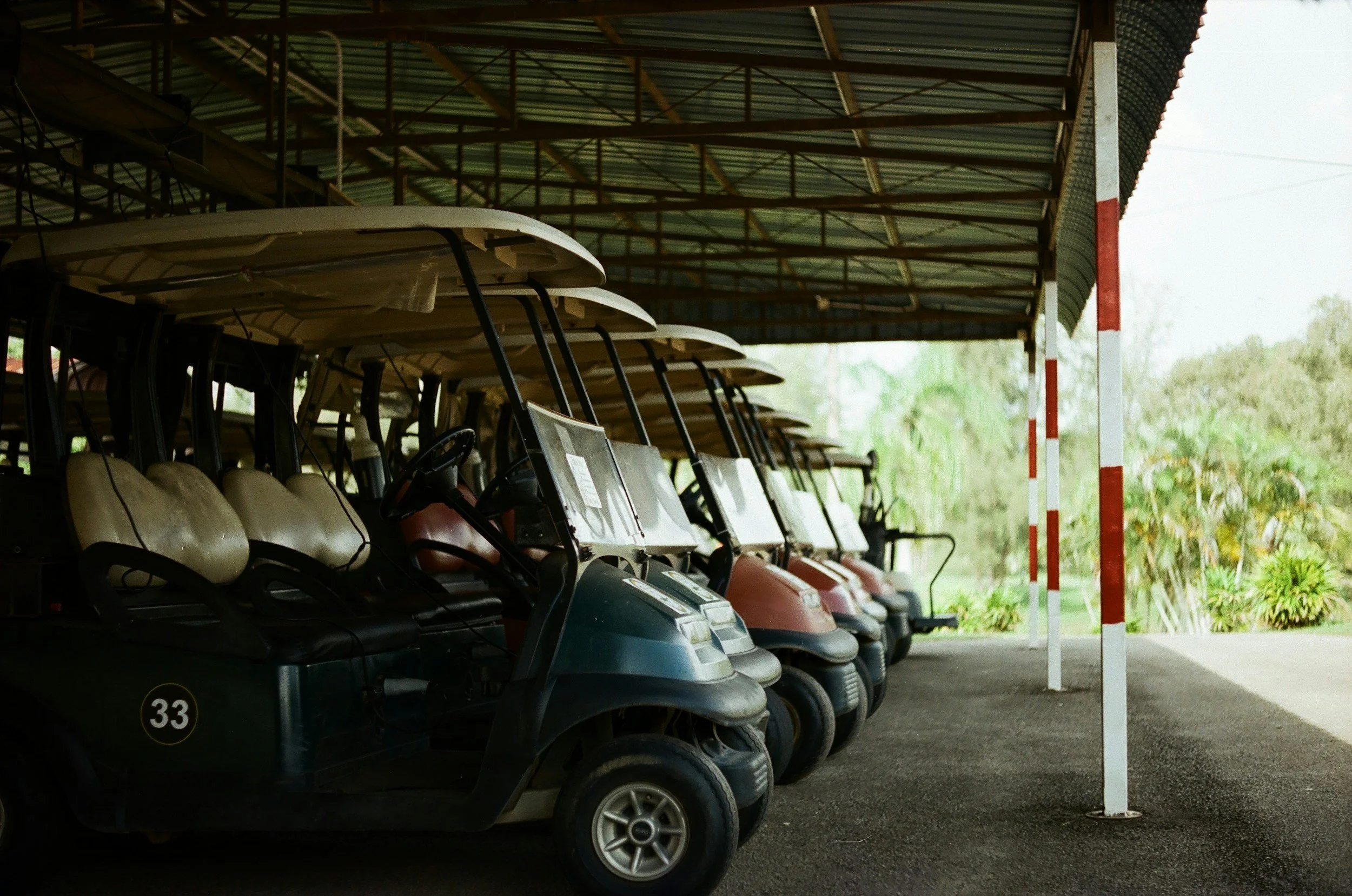 A row of parked golf carts under a covered area at a golf course, with greenery visible in the background.