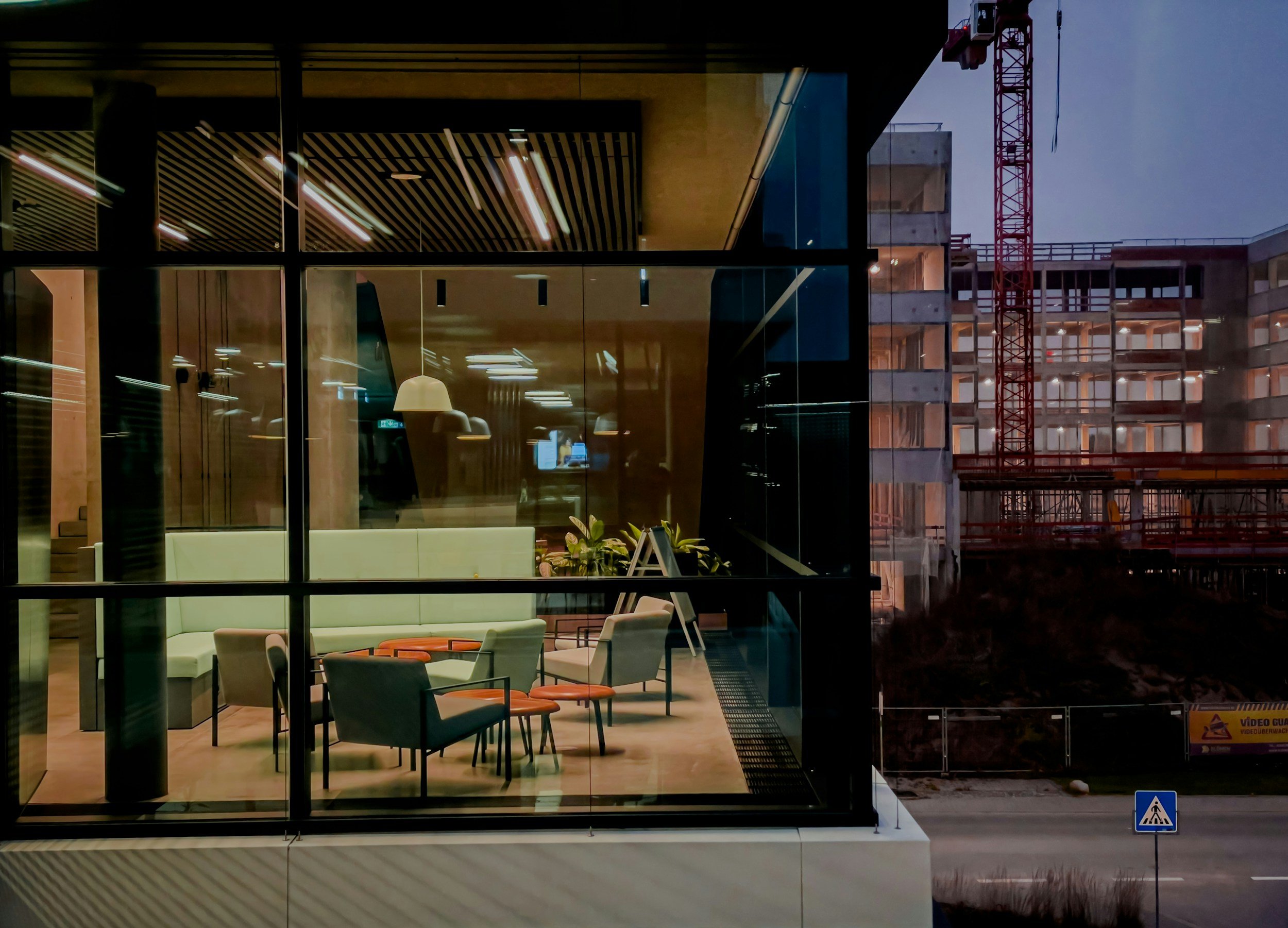 Interior view of a modern building with glass walls, showing a cozy lounge area with green sofa, chairs, and tables, illuminated by soft lighting at night, with a construction site and a crane visible in the background outside.